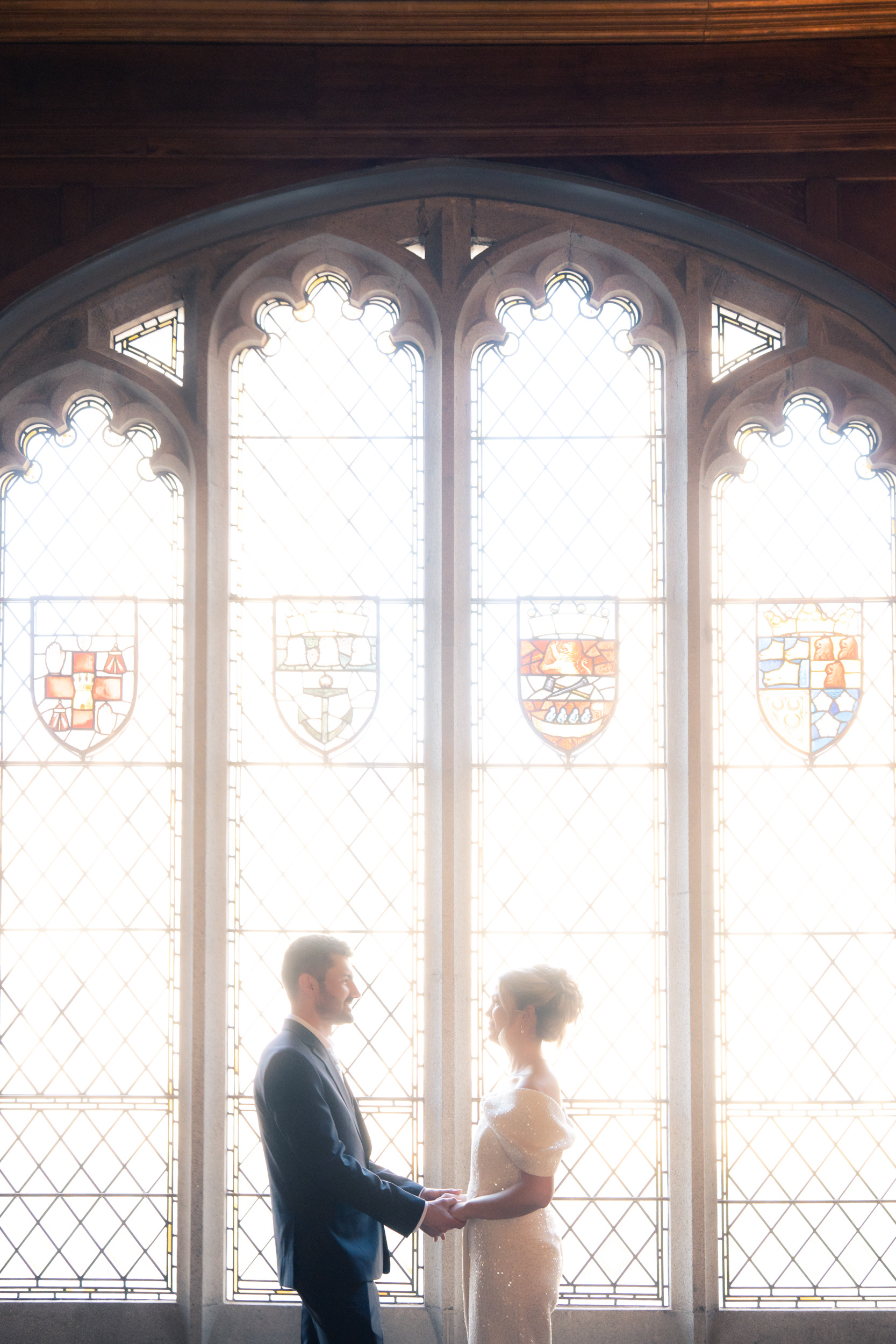 Aberdeen wedding photography: bride and groom face each other, lit by bright sunlight, in front of the stained glass window in Marischal College.