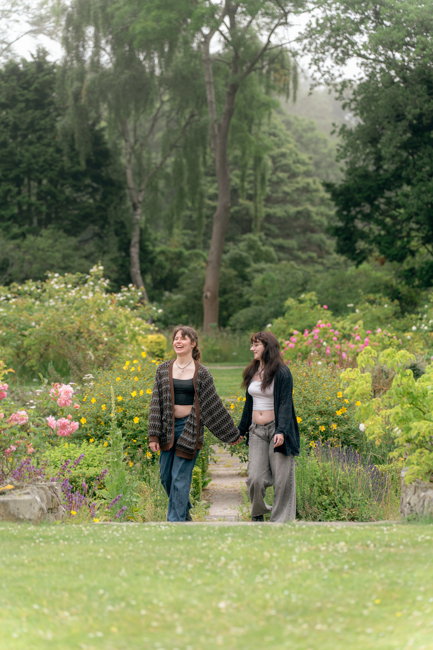 A couple (she/her and they/them) both wearing long cardigans, crop tops and jeans, hold hands and laugh as they stroll through a sunken garden during their Aberdeen photo shoot.