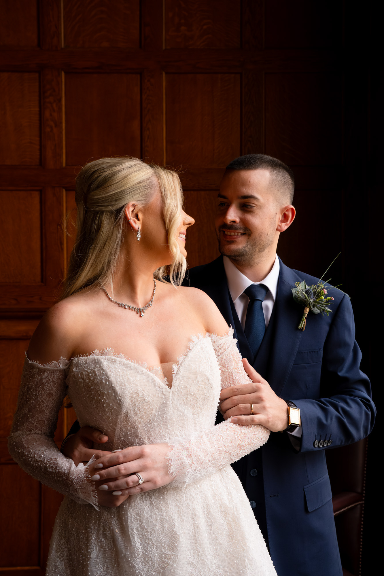 Last Minute Aberdeen Wedding Photography: A blonde bride in white mini dress and Spanish groom in blue suit gaze at each other, side lit, with wood panelled wall as backdrop.