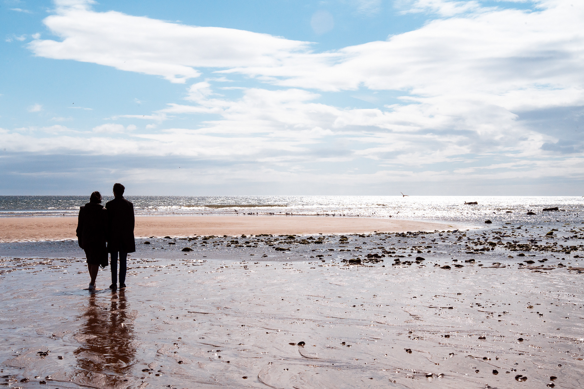 A silhouetted couple look out to sea at low tide on Aberdeen Beach. It's a cold day but the sky is blue.