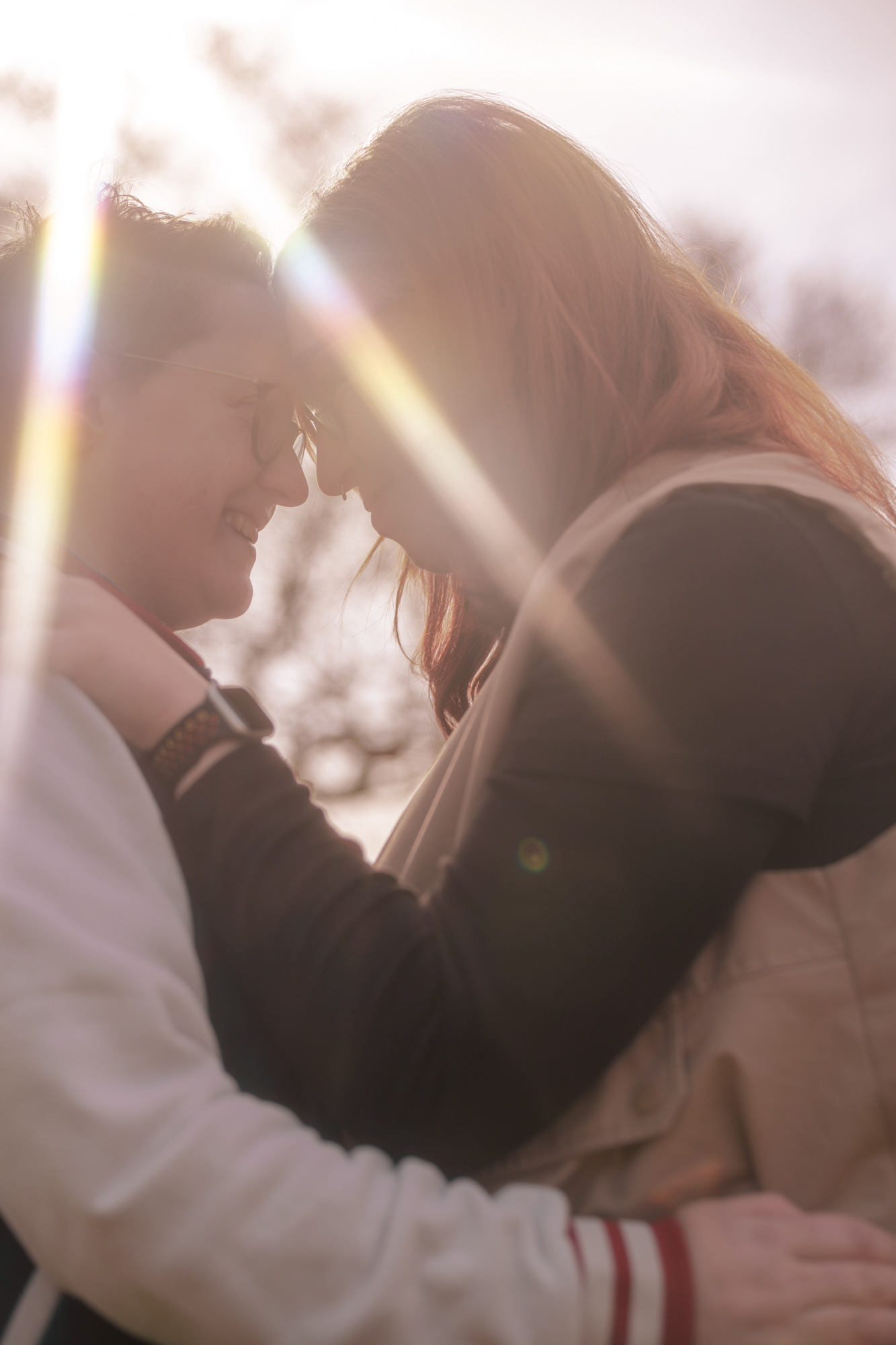 Close up of a couple (she/her and they/them) leaning in for a kiss, rainbow sunburst shining above them.