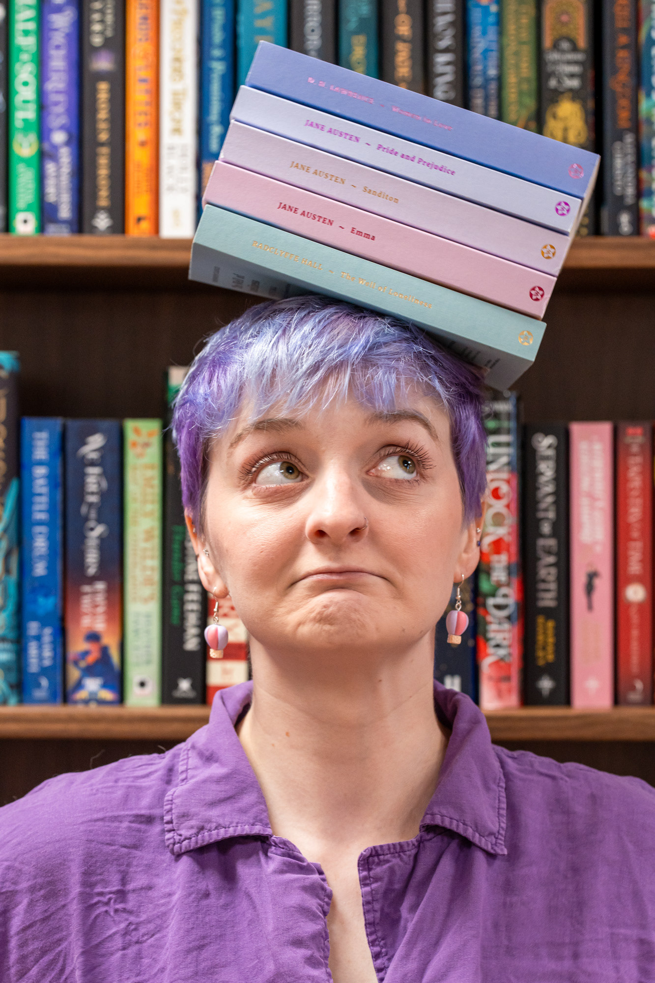 Somerville Books Bookshop owner, Christie, a white woman with short purple hair and amazing bird, cat and floral tattoos looks up to one side as a pile of pastel Jane Austen books slides off her head. Aberdeen main character photo shoot.