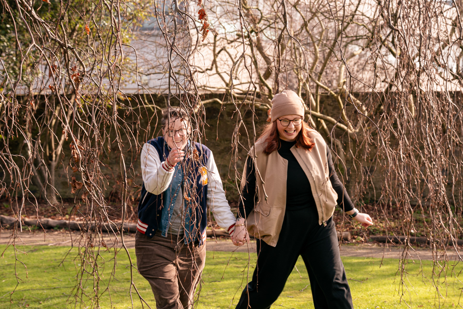 A queer couple (she/her and they/them) laugh as they run through the branches of a low-hanging tree in Aberdeen