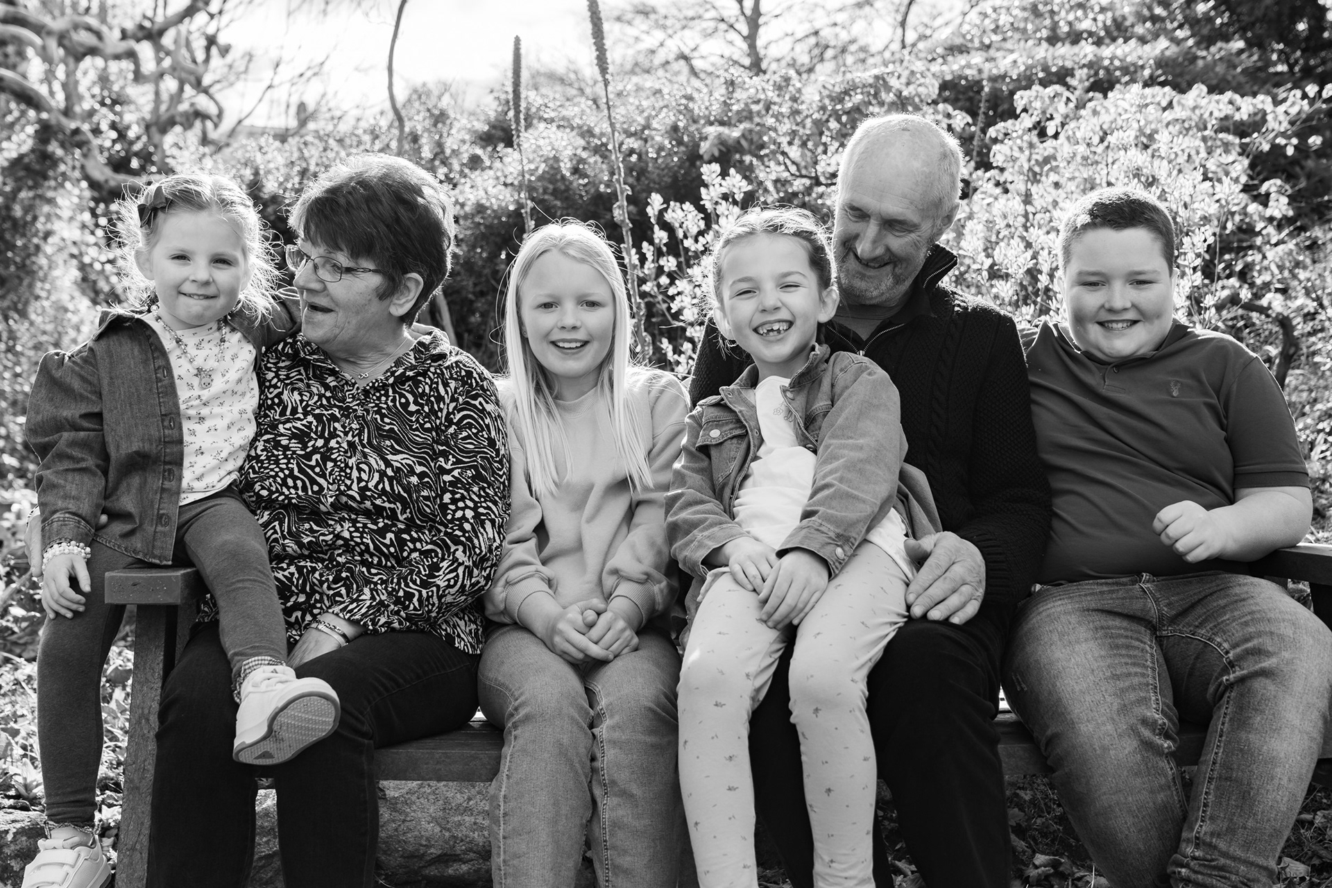 A black and white image by Aberdeen family photographer, Windswept Stories, shows two grandparents and four grandchildren - three girls and a boy - sitting on a bench together, giggling.