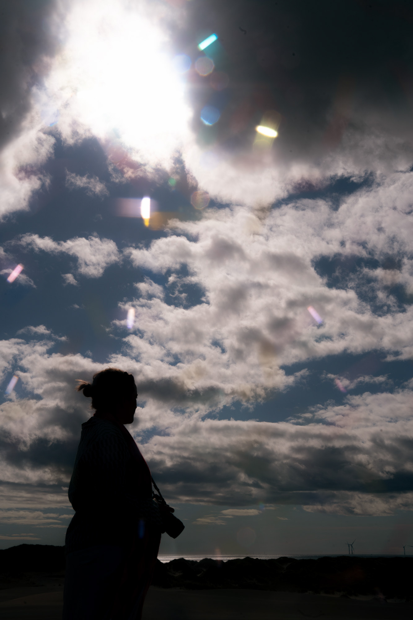 A woman's head and shouders are silhouetted against a cloudy sky, with a bright spot where the sun is trying to come through, and colourful flares of light bouncing around her.