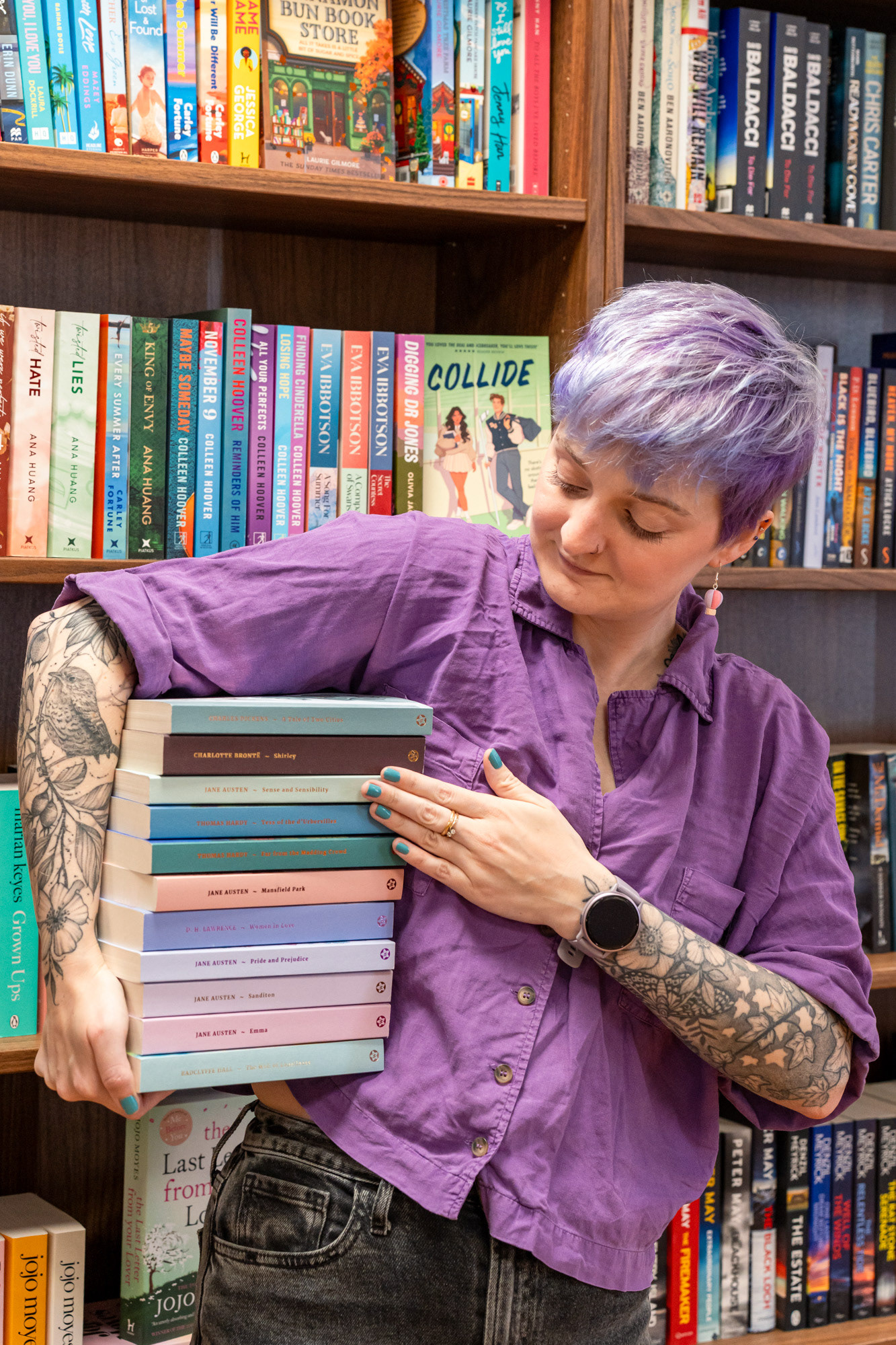 Aberdeen bookshop branding photography: A white woman with purple hair and floral tattoos stands in front of shelves of colourful books, a stack of pastel classics under one arm.