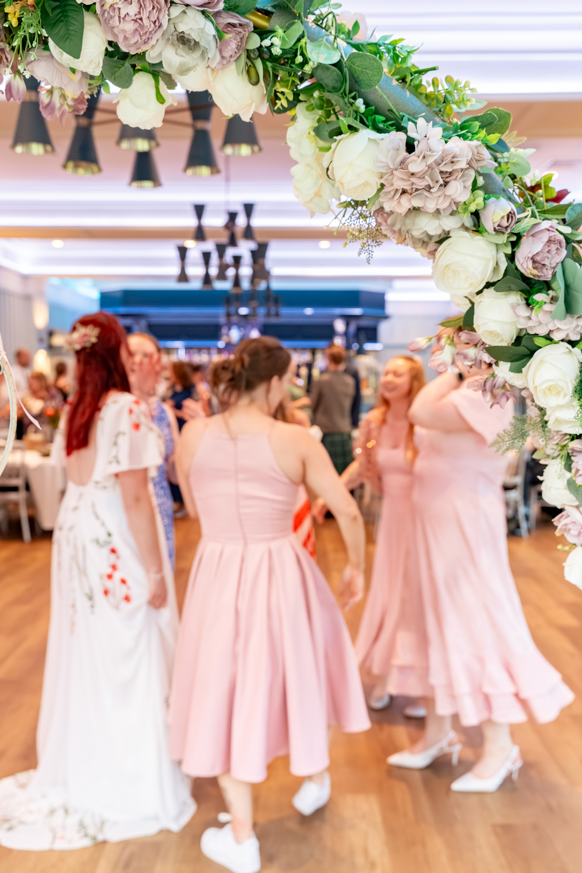 A bride in a floral dress and her bridesmaids in various shades of pink are shown through a floral arch, dancing at her Aberdeen wedding
