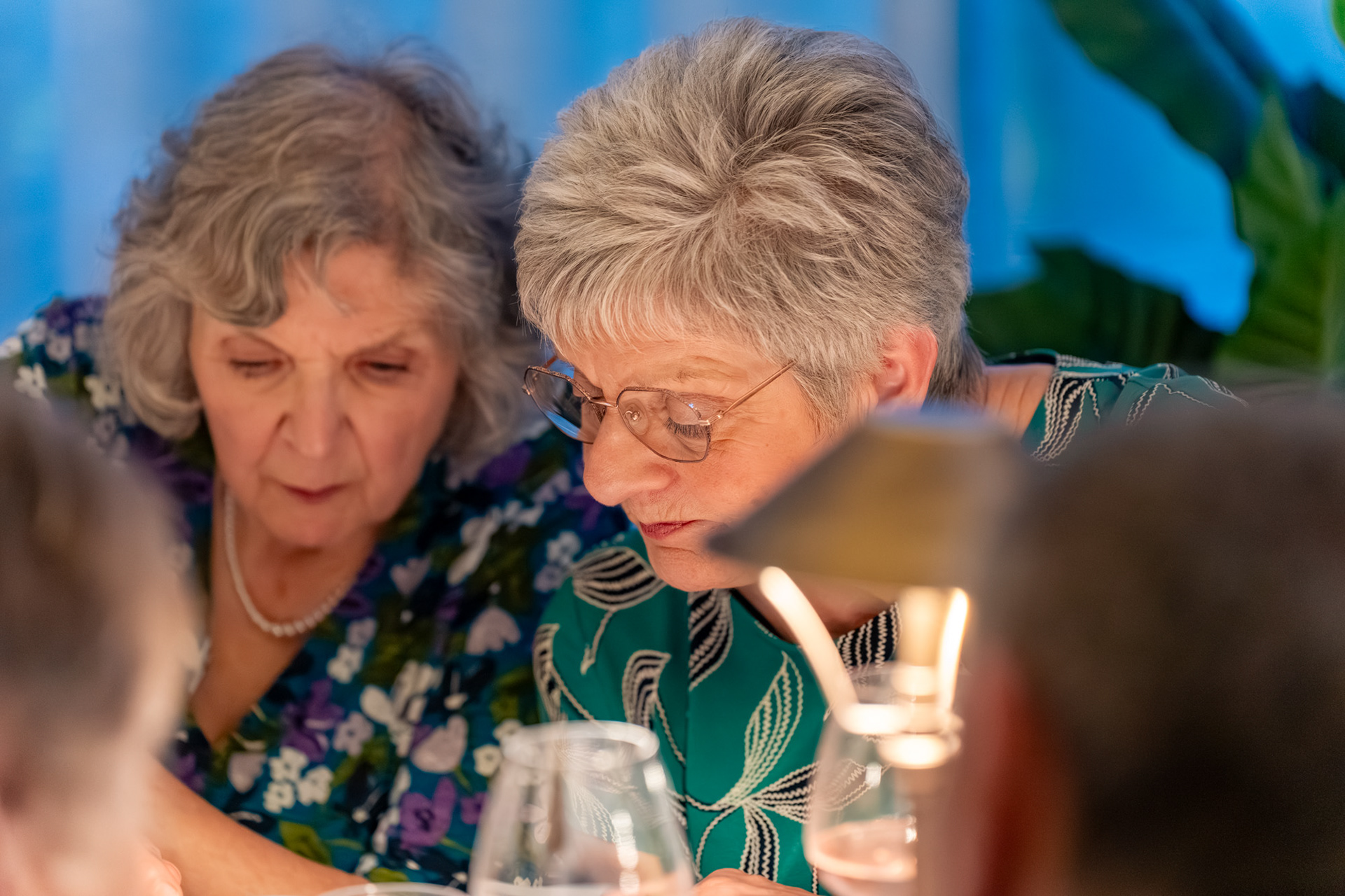 Two grey-haired women in floral dresses lean their heads close together in thought.