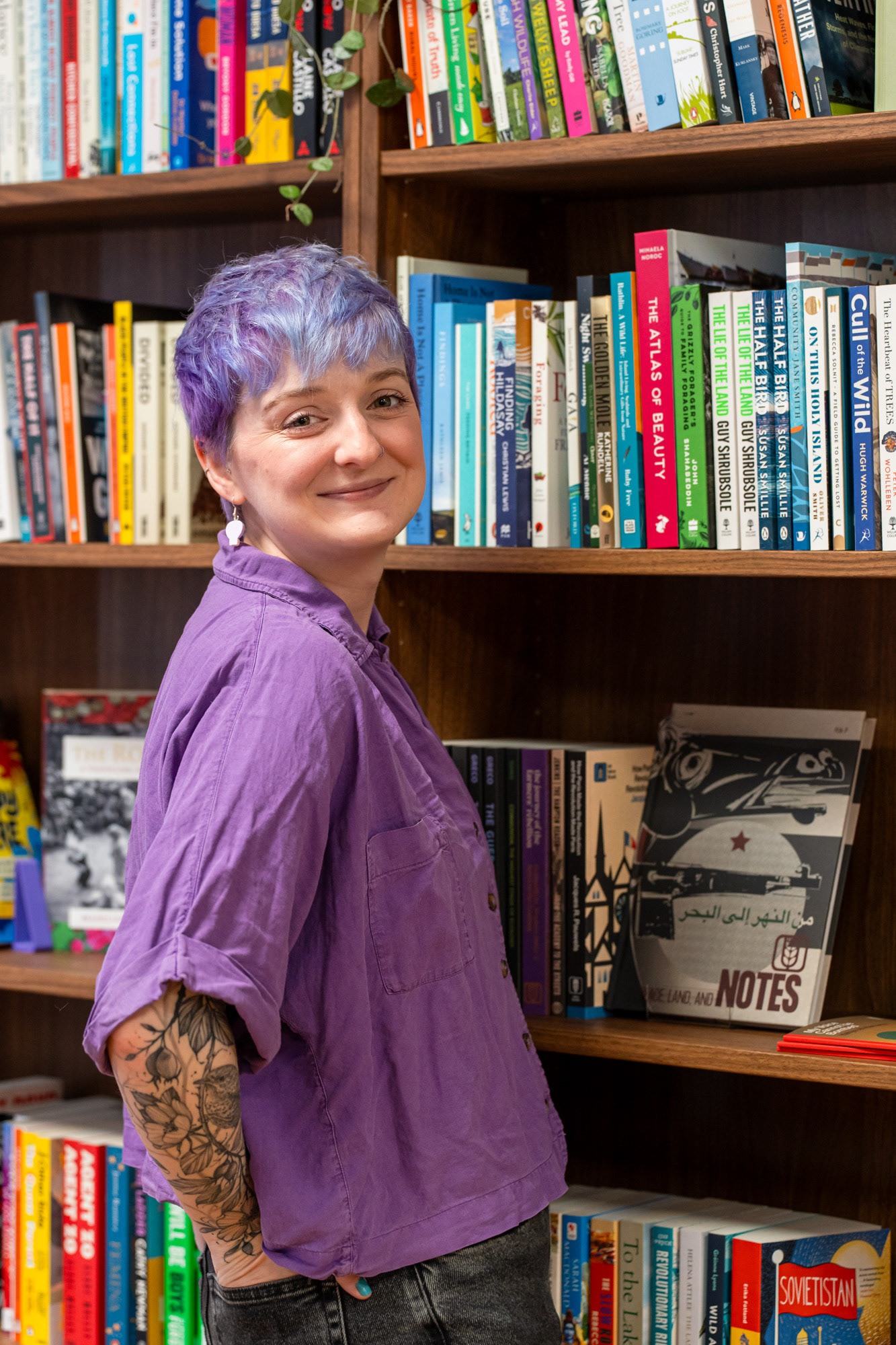 Somerville Books Bookshop owner, Christie, a white woman with short purple hair and amazing bird, cat and floral tattoos looks back over her shoulder and smiles at the camera; the backdrop is shelves of colourful books.