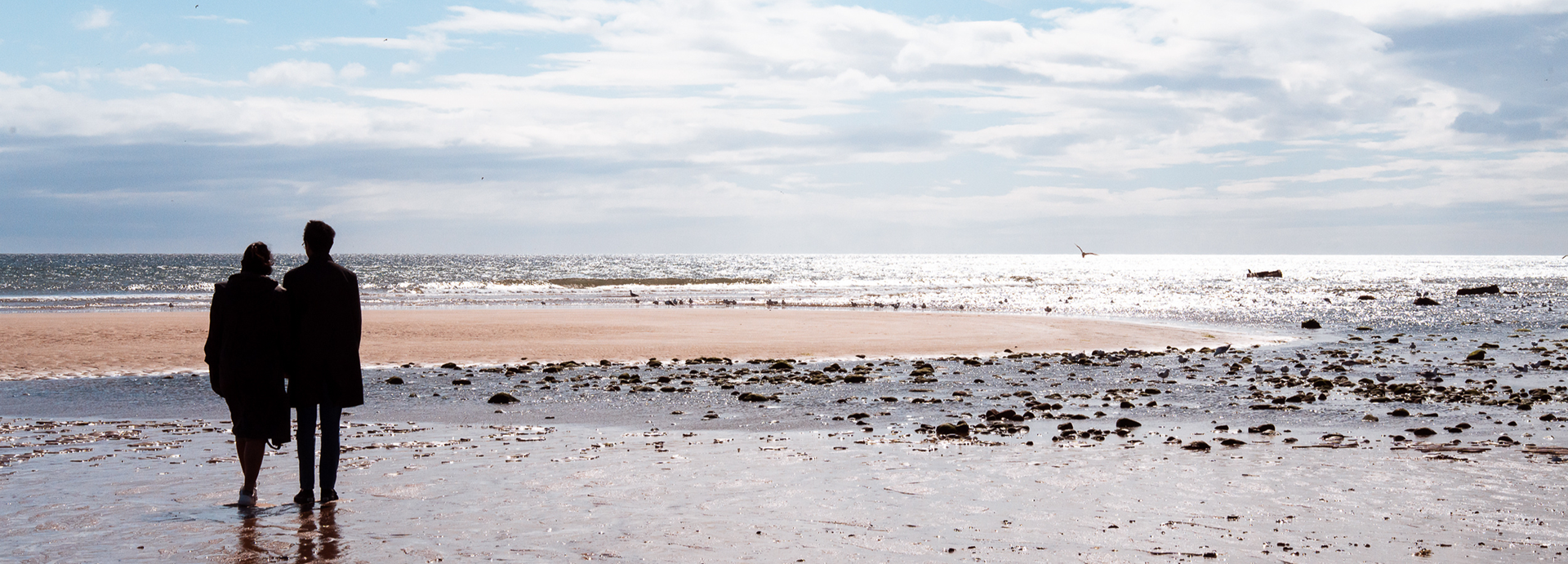 During their Aberdeen engagement photo session, a couple (one female, one male) are silhouetted as they stand together, gazing out to sea. The wate shines with sunlight around them.