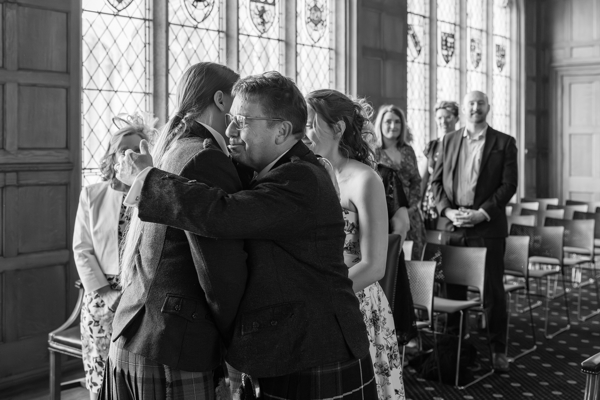 Father of the bride hugs the groom at the end of the aisle as the bride and guests look on.