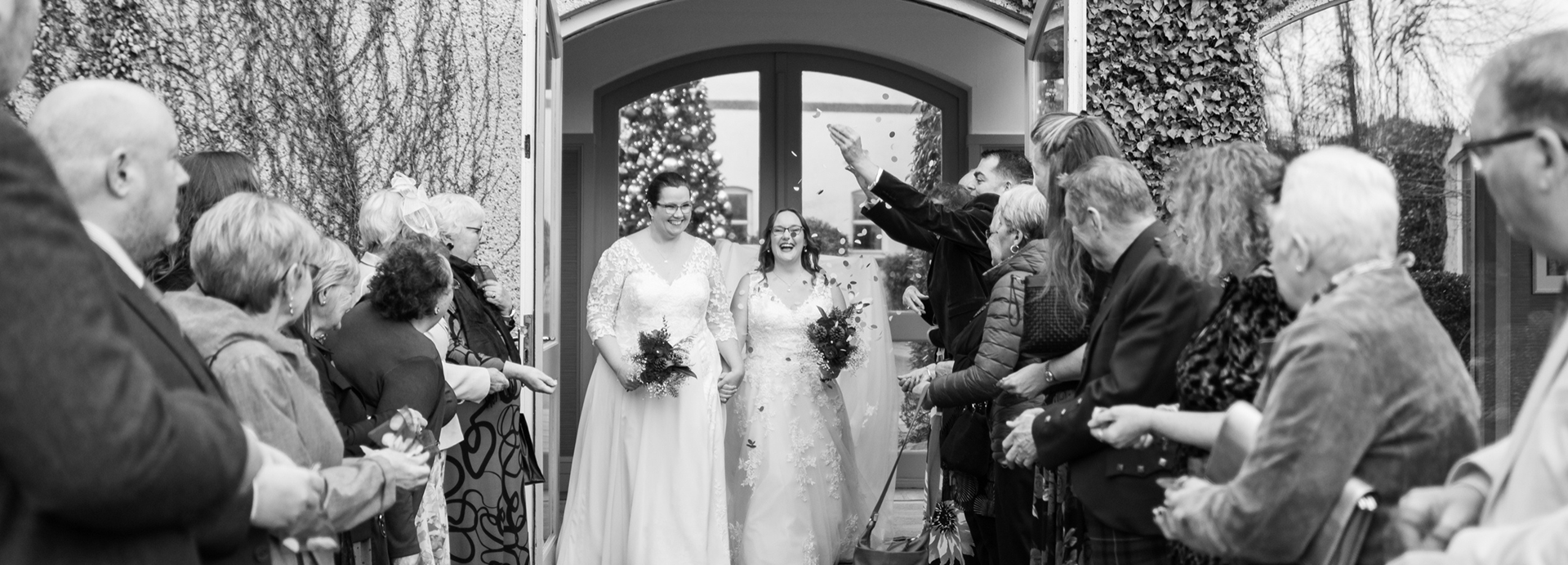 A black and white image of two brides in white dresses being showered by confetti by their assembled guests, at a same-sex lesbian wedding in Aberdeen.