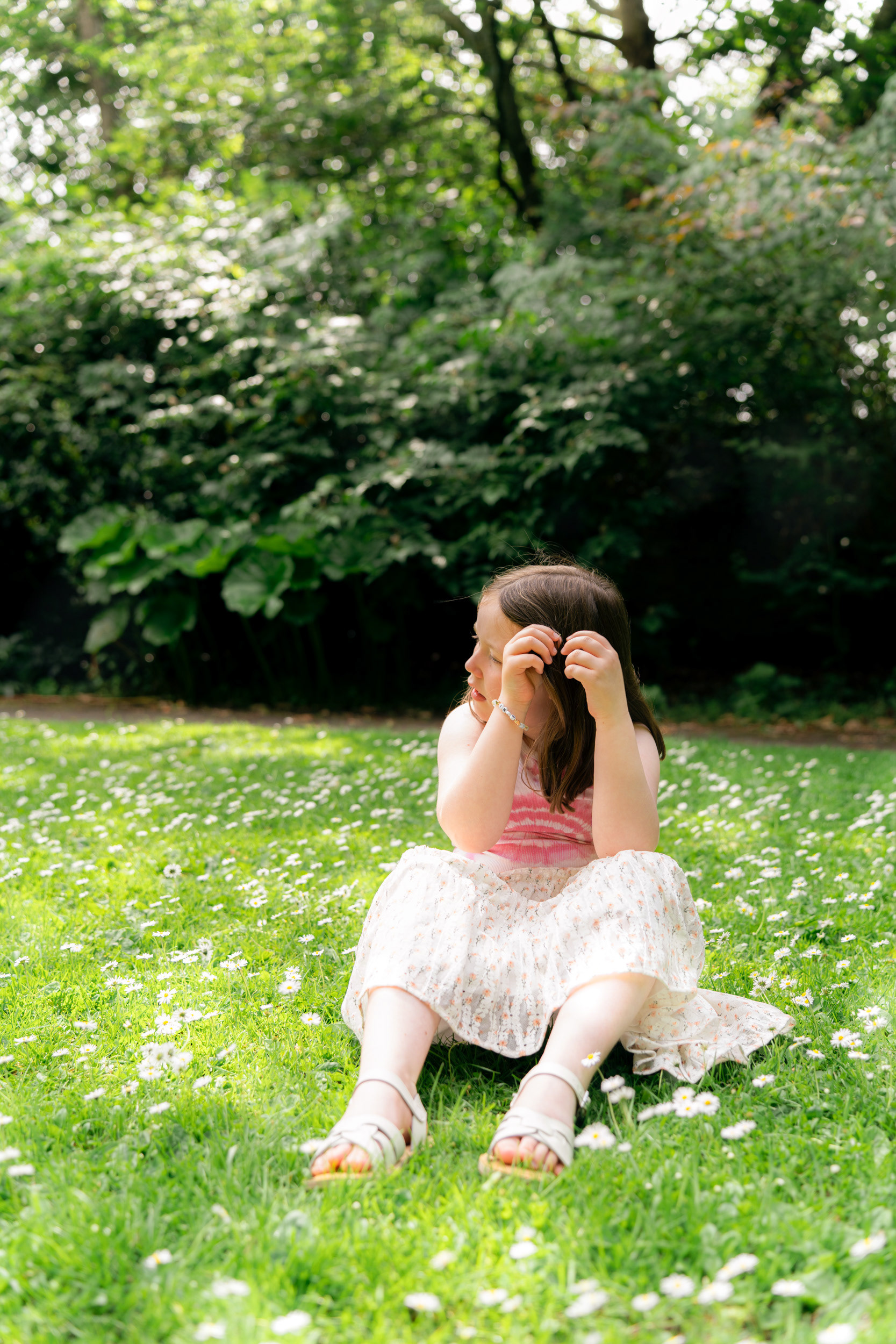 In an Aberdeen park, a 7 year old girl in sandals, vest and floral skirt, sits amongst the daisies, trying to tuck one behind her ear.