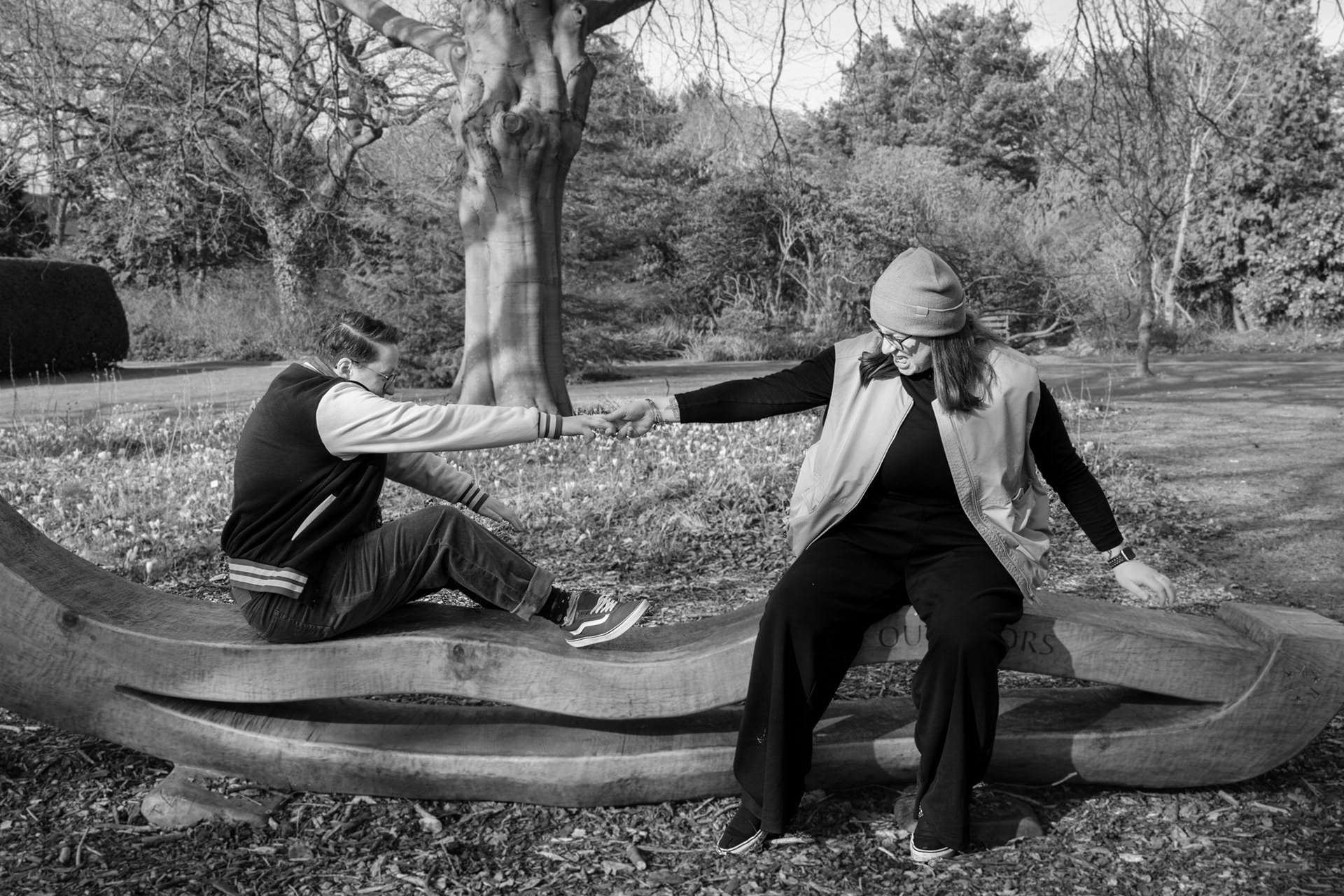 A queer couple (she/her and they/them) mess around, pushing and pulling on an ornamental bench in Old Aberdeen.