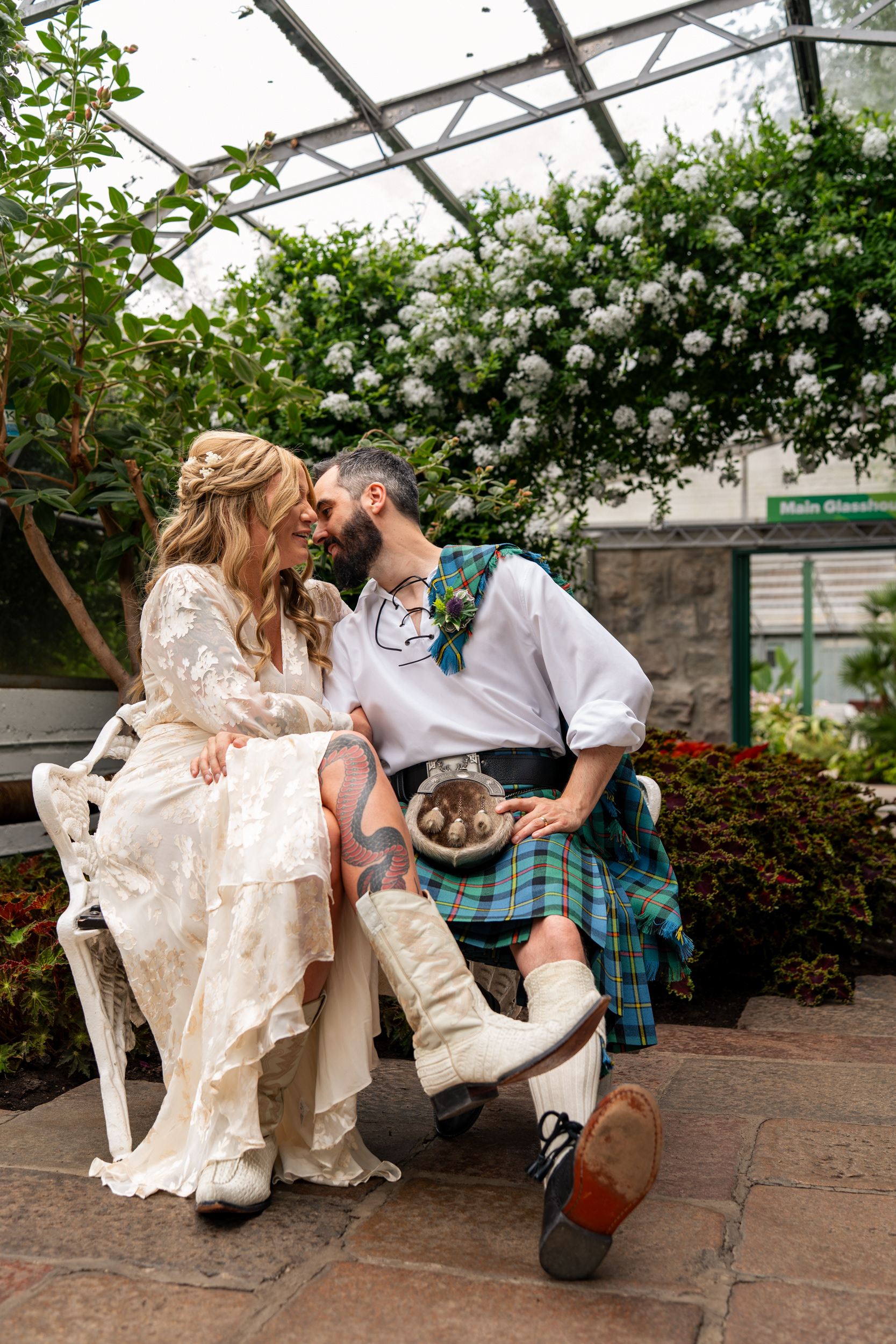 Aberdeen Duthie Park Winter Gardens wedding: Bride in boho ivory dress and cowboy boots and groom in blue and green lounge on a white metal bench under an arch of white flowers.