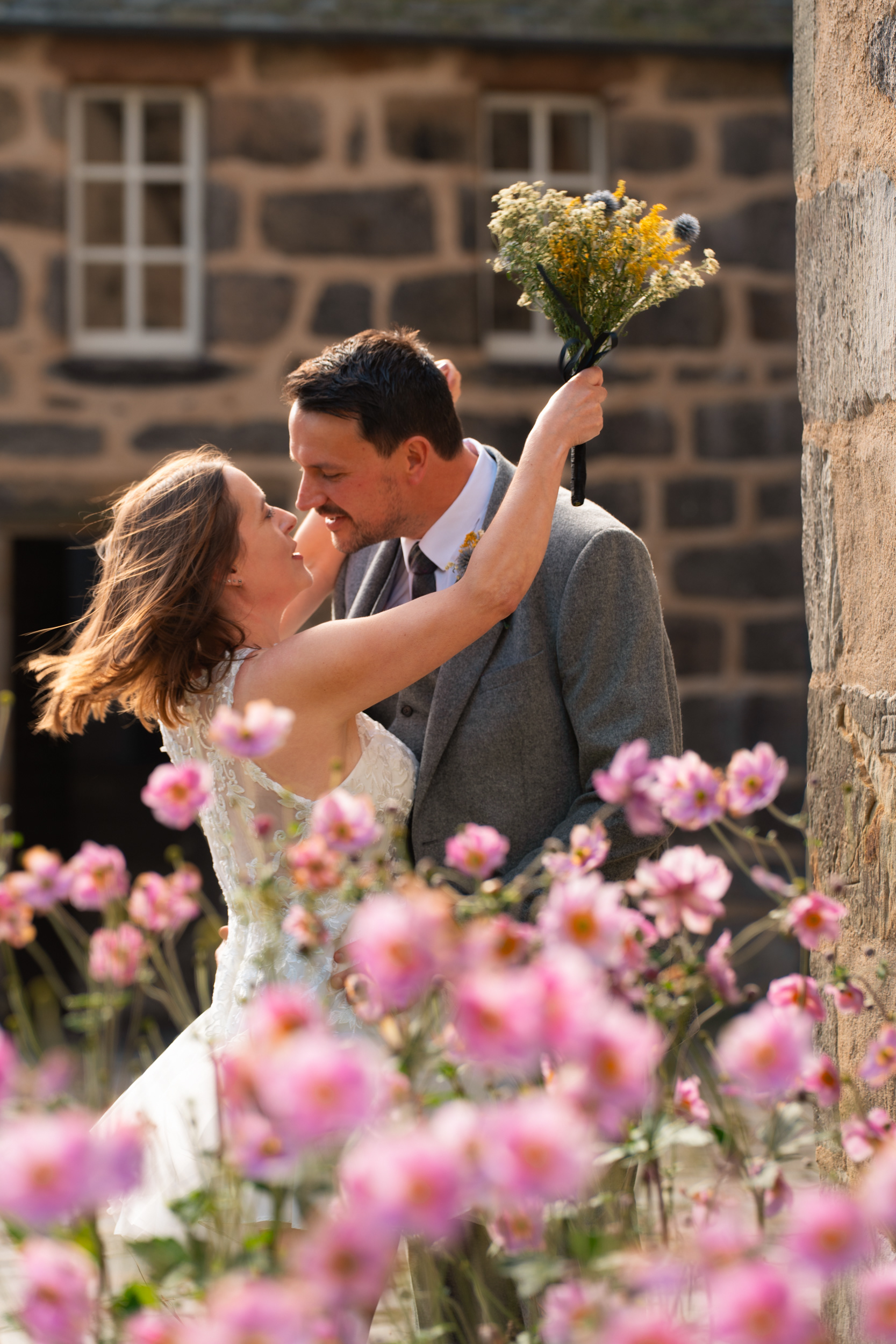 Marischal College wedding, Aberdeen city centre - Bride in white and groom in grey suit lean in for a hug, hair caught by the wind, pink anemones dancing in front of them.