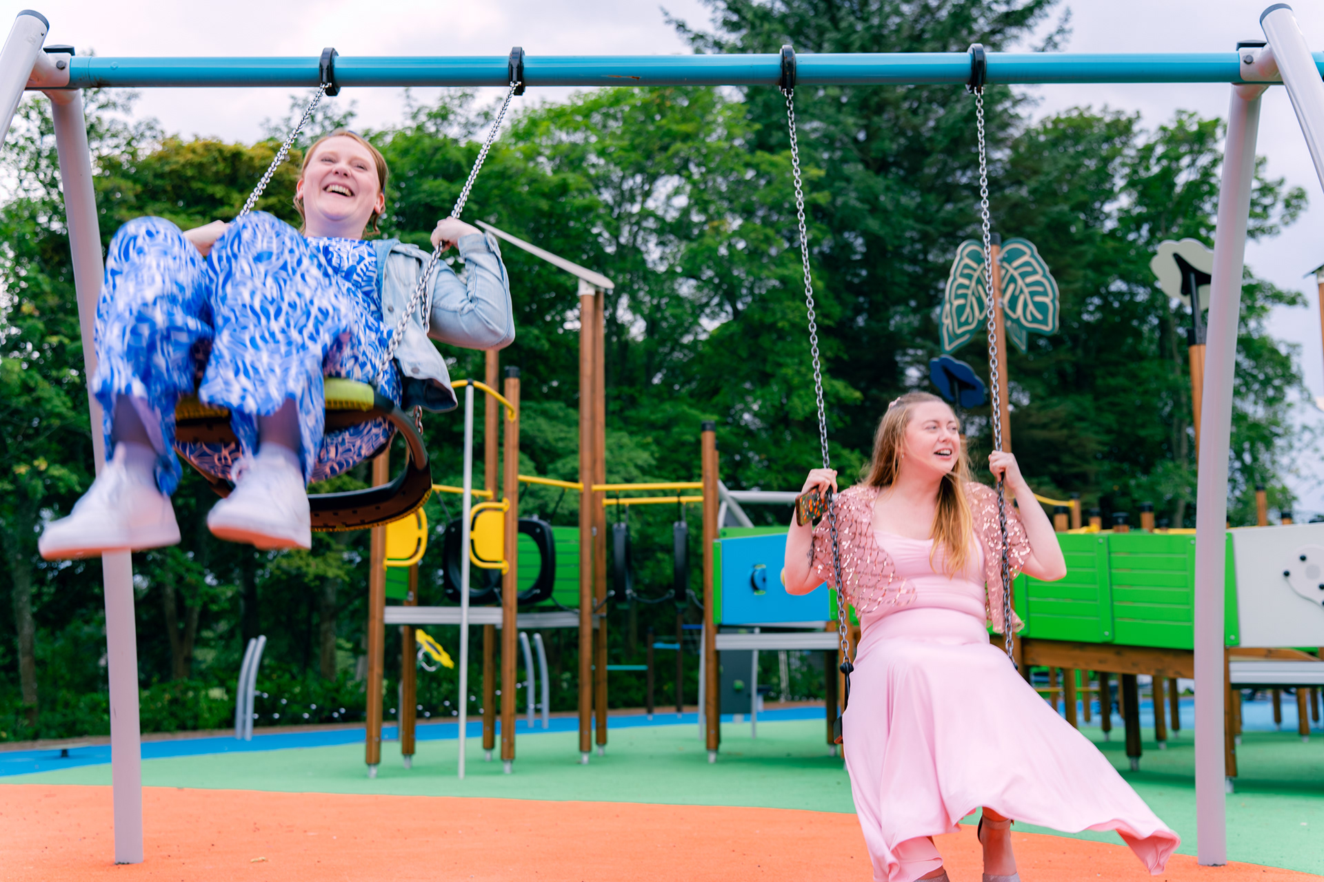 Two young women - one in a blue jumpsuit and one in a pink bridesmaid dress and cape - laugh on the swings at Hazlehead during an Aberdeen wedding.