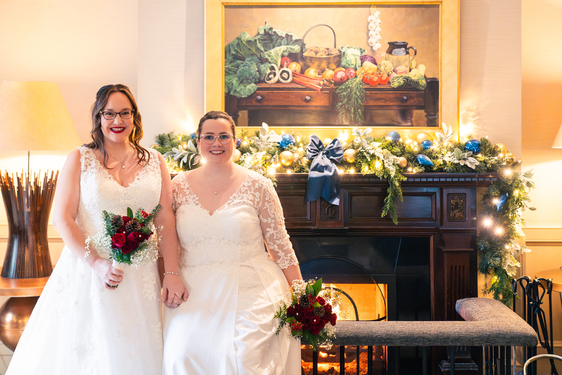 Two brides in white dresses hold hands, sitting in front of a roaring fire decorated for Christmas in navy and gold.