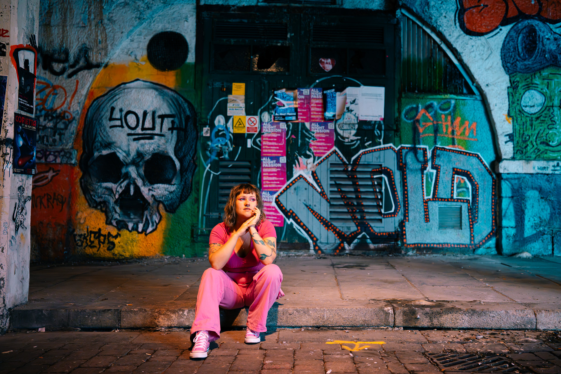 A young queer woman, dressed in pink top, pink trousers and pink Converse, sits on a kerb with a heavily graffitied wall behind her. Her chin is resting on one hand and she looks off to one side, pensively.