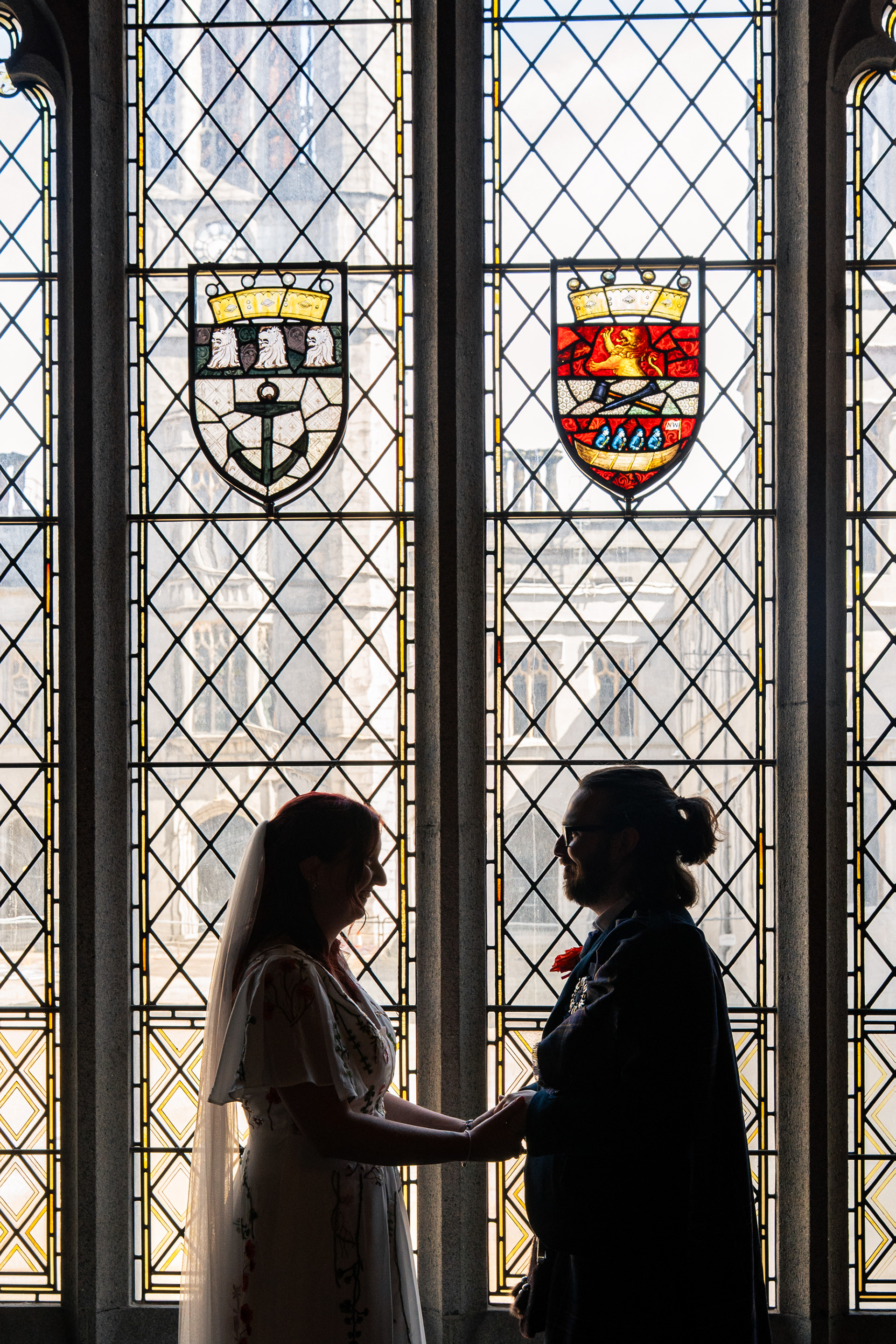 Bride and groom are silhouetted against the stained glass window in the Grant Room of Marischal College after their wedding ceremony. They are holding hands and appear to be smiling at each other.