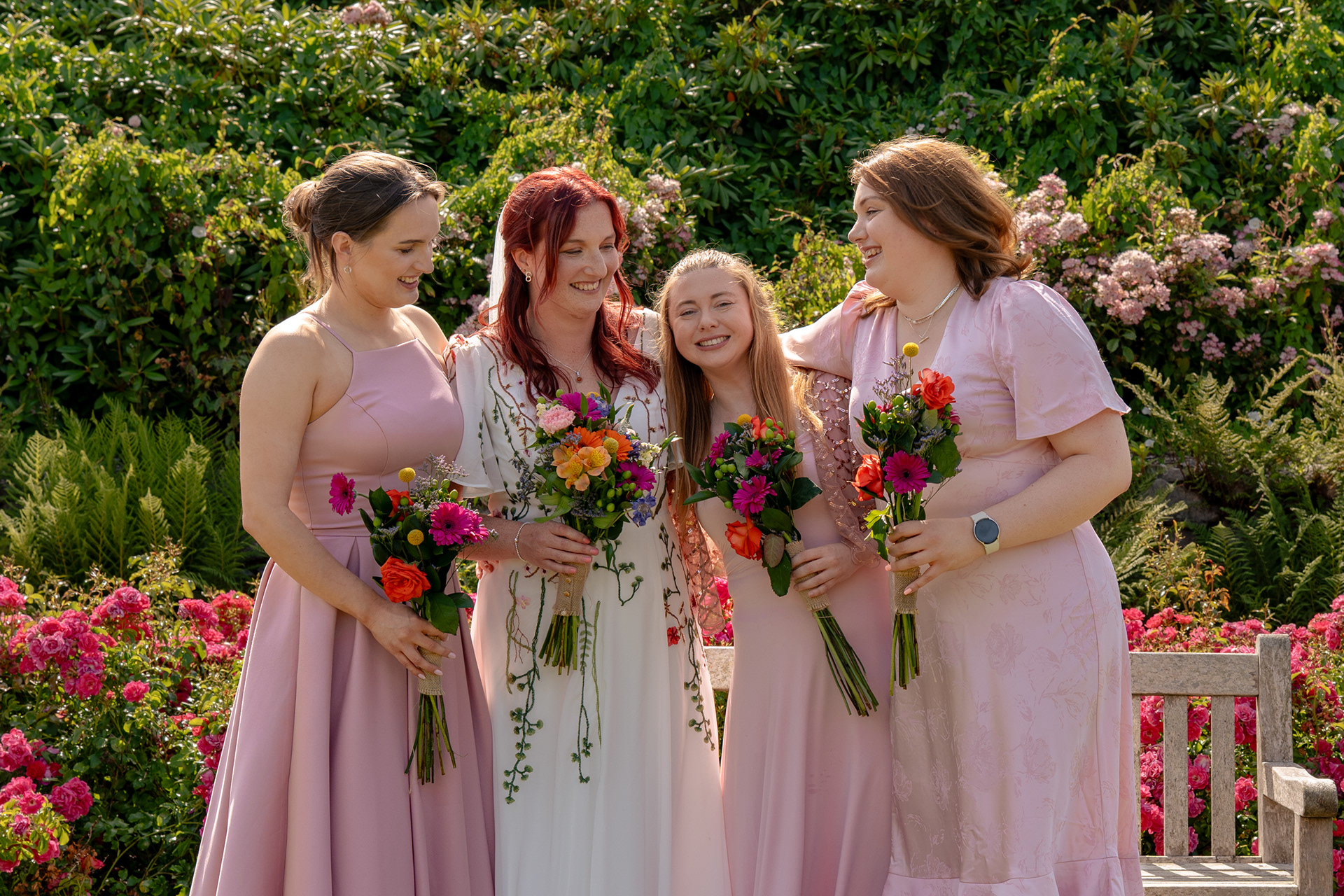 A bride in a floral dress and three bridesmaids in different pastel pink dresses laugh together, all holding colourful bouquets, in the Hazlehead Park rose garden.