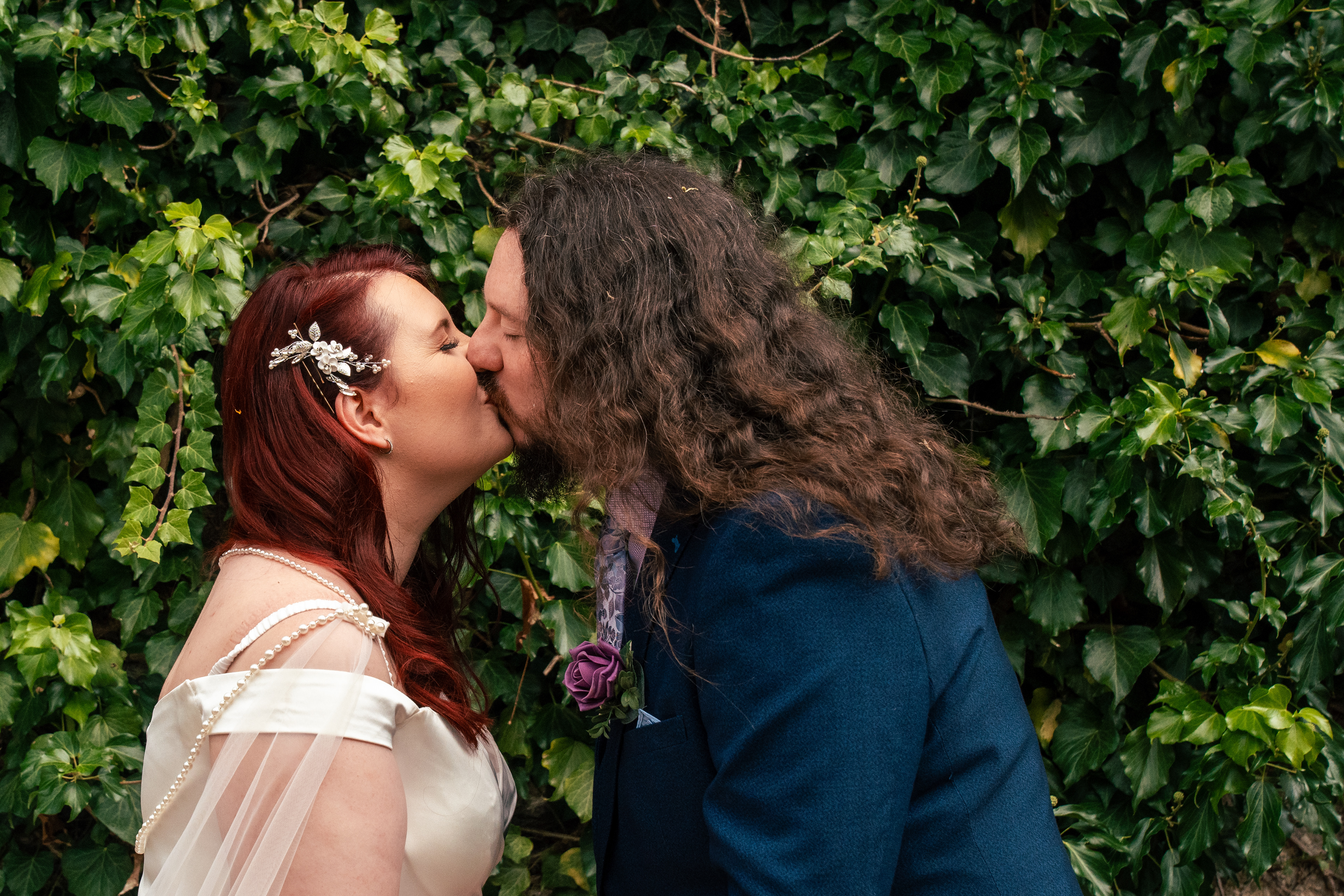 Red-haired bride in ivory dress and long-haired groom in navy suit kiss against a backdrop of ivy in Aberdeen's Winter Gardens.