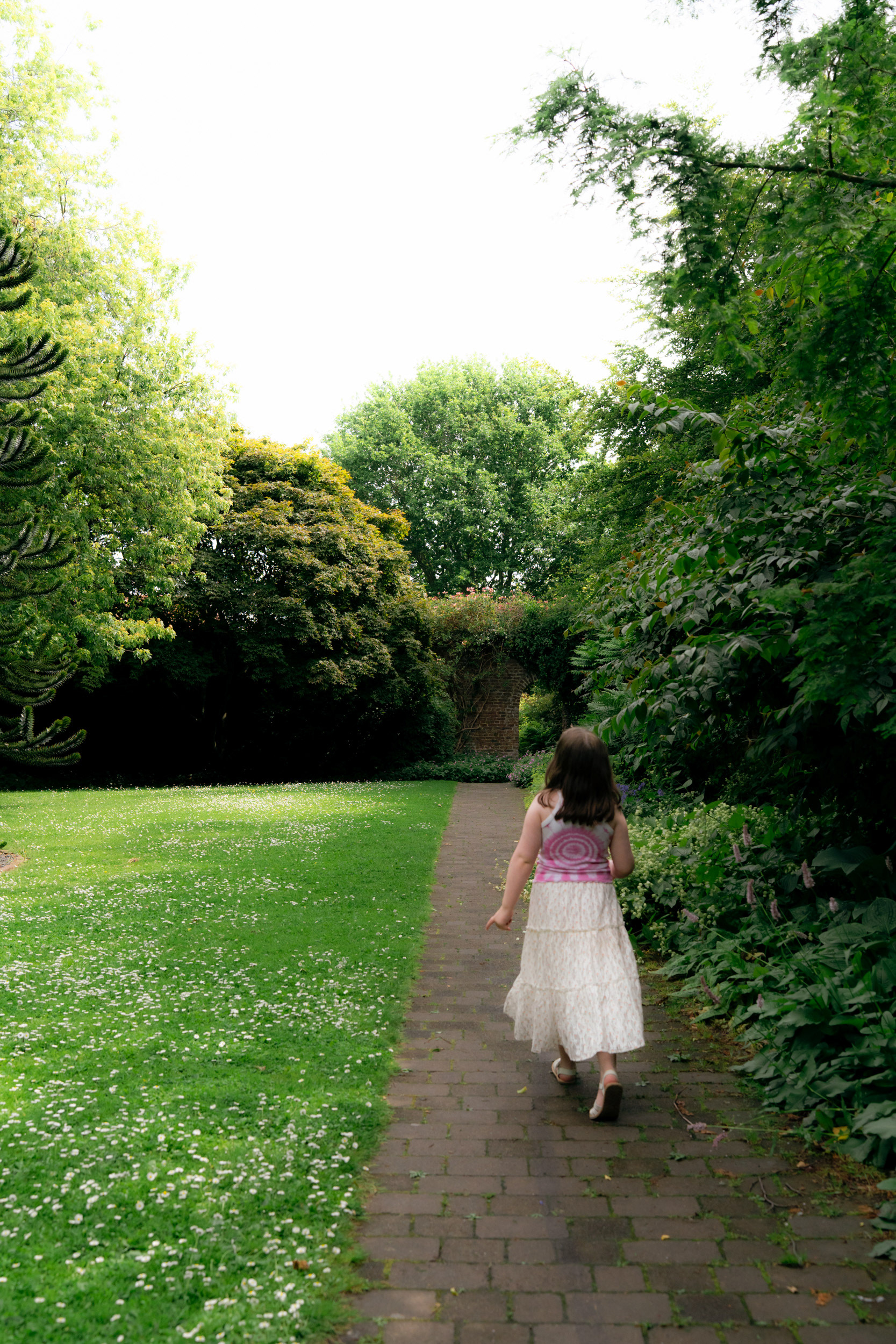 A young girl in a pink tie-dyed vest and floral skirt walks away from the camera on a sunny day, in an Aberdeen walled garden.