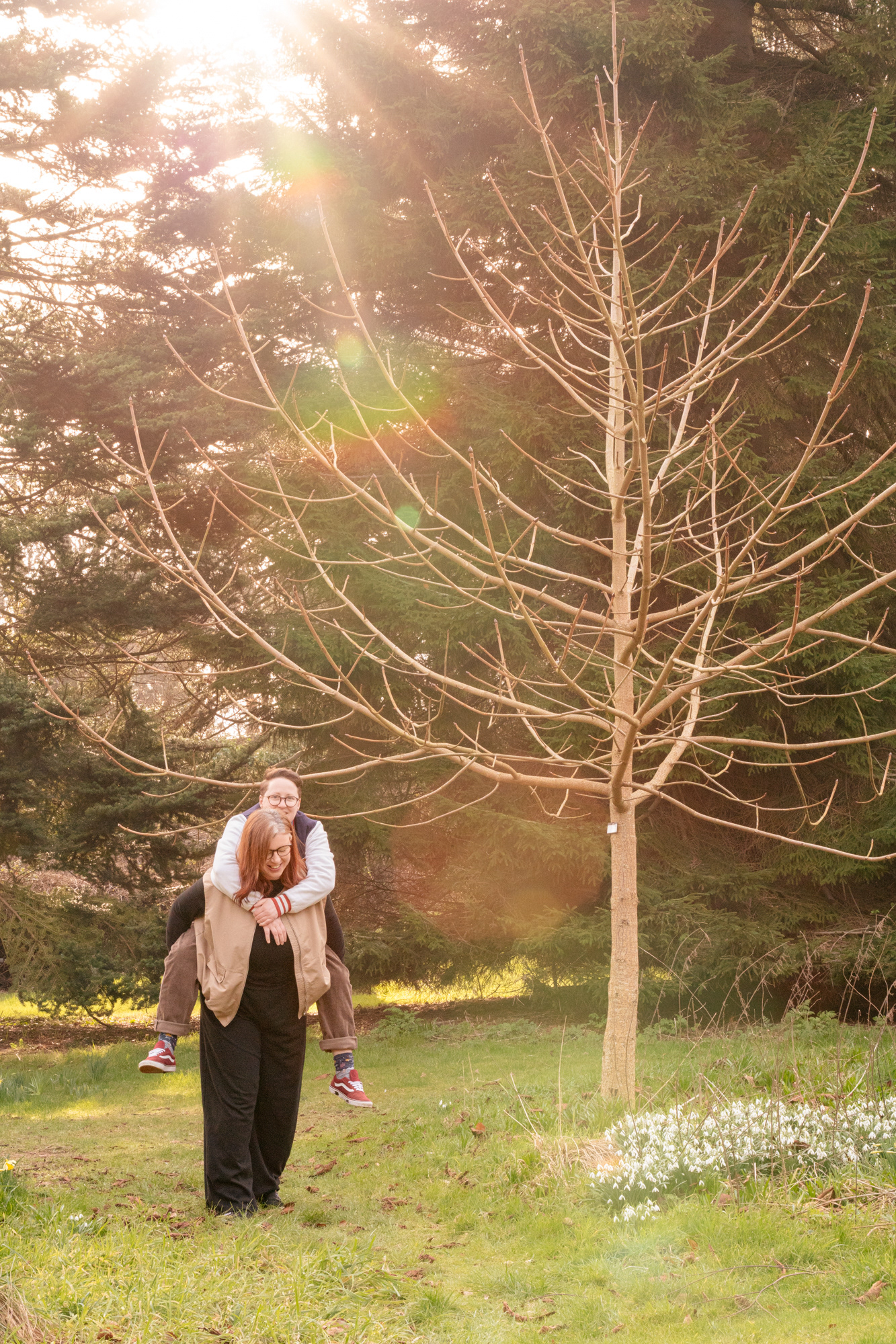 A lesbian (she/her) gives her partner (they/them) a piggy back past a patch of snowdrops, rainbow beams of sunlight shining above them, during a queer couple's photo shoot by Aberdeen wedding photographer, Windswept Stories
