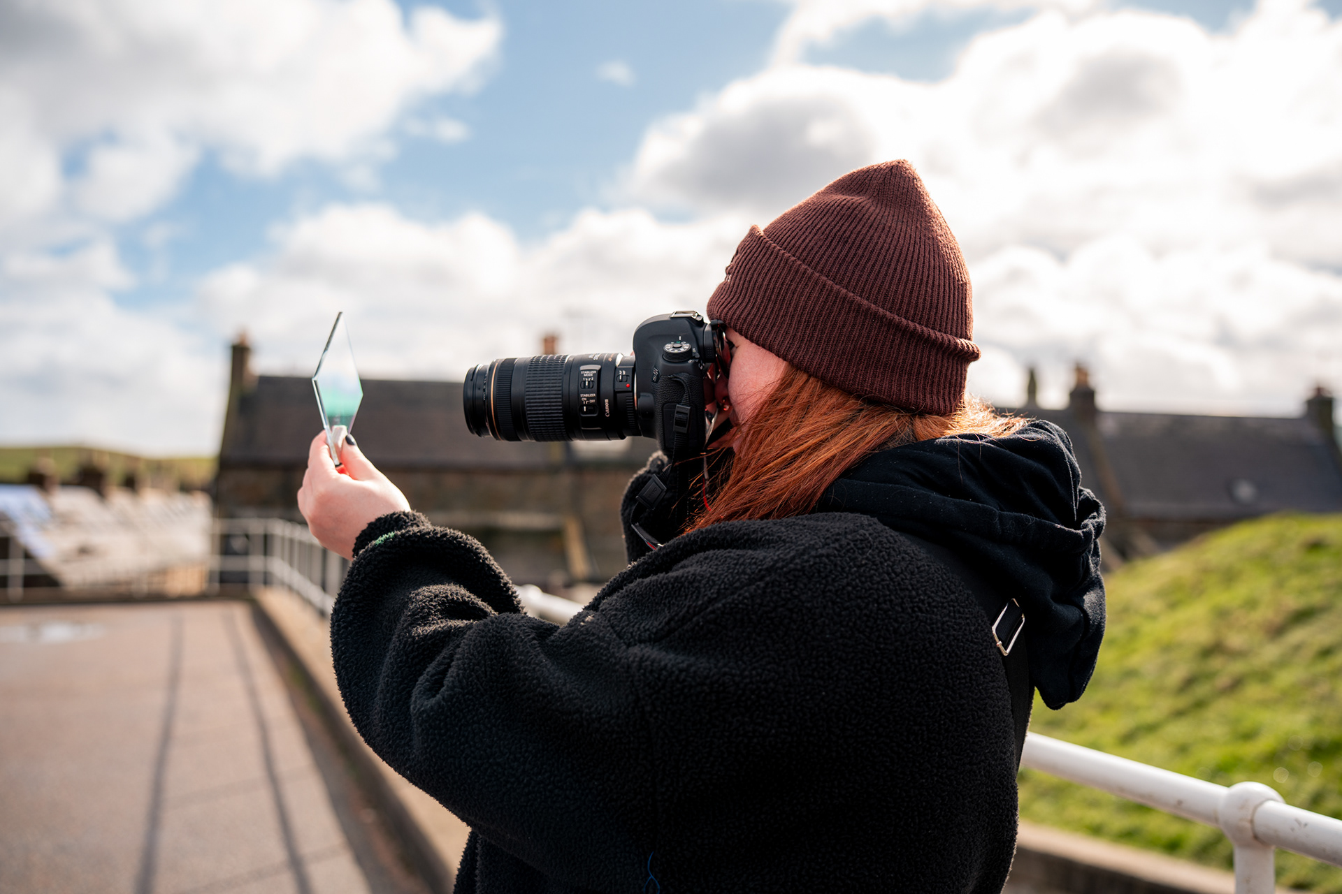 A woman in dark clothes and beanie hat takes a photo using a DSLR pointing through a sheet of iridescent glass.