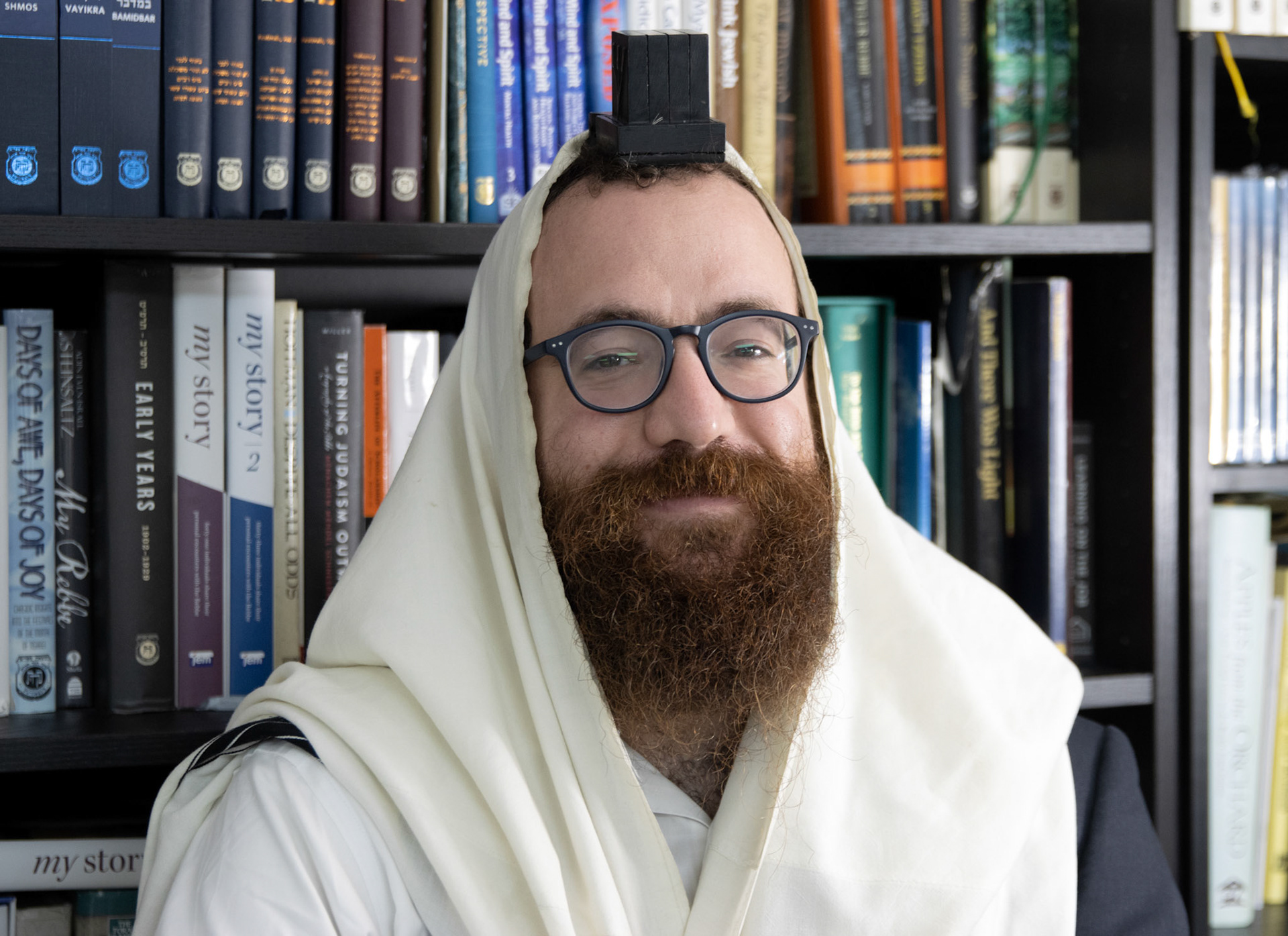 Berel Gurevitch, 33, stands for a picture in front of the bookshelves at the University of Oregon brand of chabad where he is the Rabbi, on Monday, May 8, 2023, in Eugene, OR. He was wearing his tallit and tefillin in preperation for his daily prayers. (Photo by John Ofstedal)