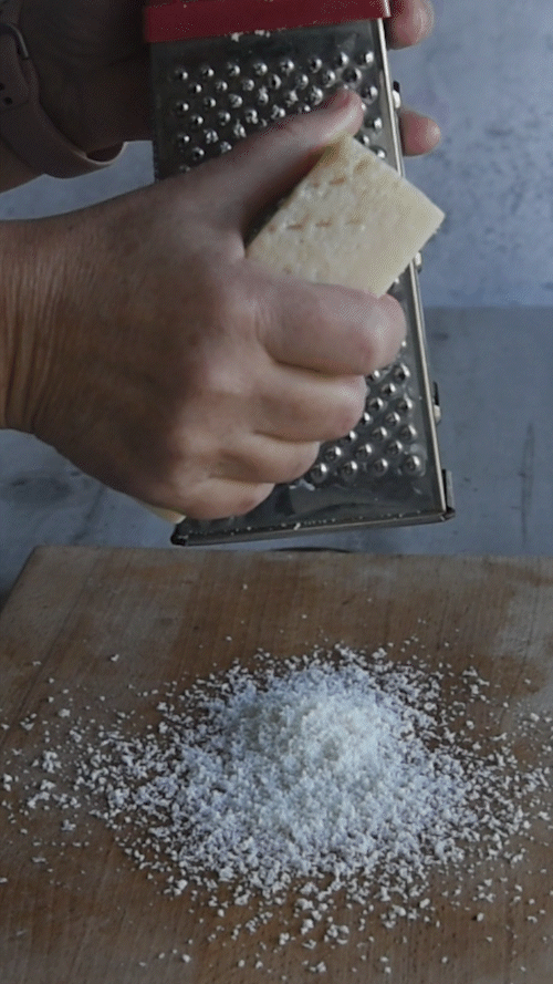 Primo piano di una mano che grattugia un pezzo di formaggio a pasta dura su un tagliere di legno, con le scaglie che si accumulano in un loop continuo.
