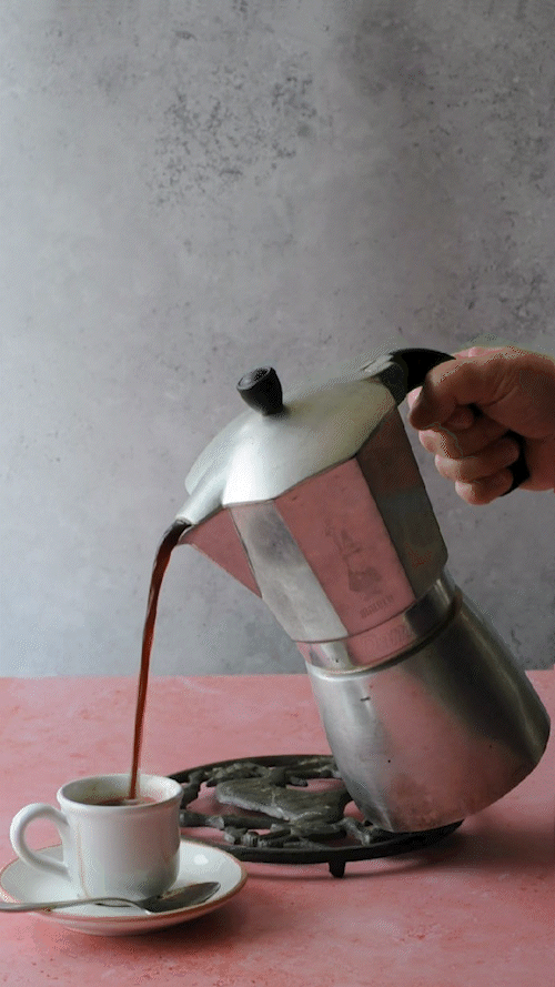 Una Moka che versa caffè espresso fresco in una tazzina di ceramica bianca su una superficie rosa.