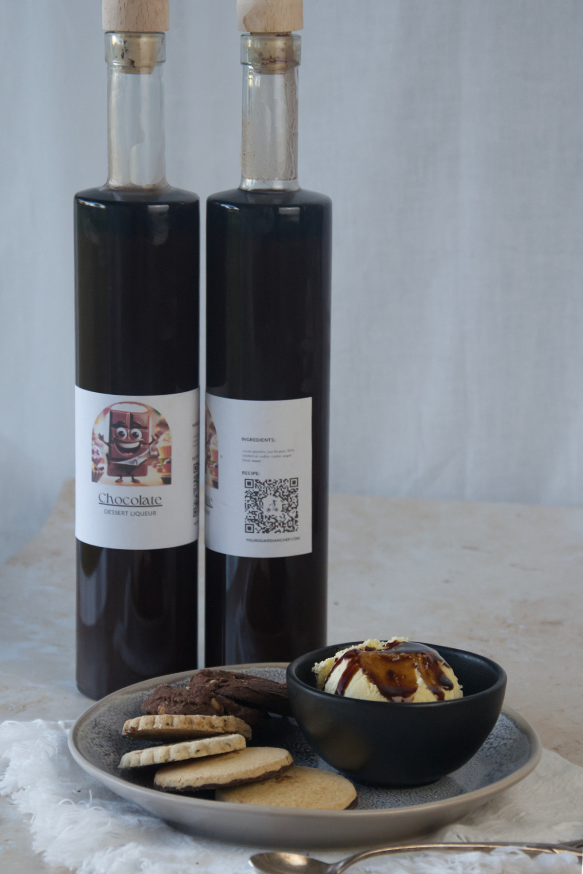 Due bottiglie alte di liquore al cioccolato fondente dietro una coppetta di gelato alla vaniglia con guarnizione al cioccolato e biscotti assortiti su superficie neutra.