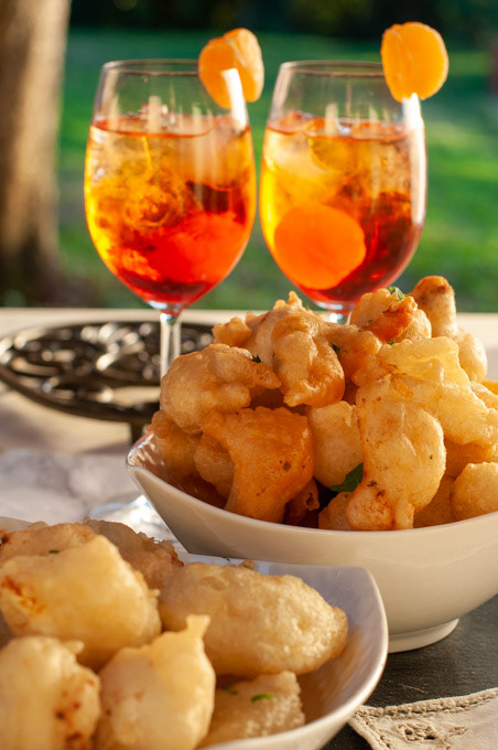 Crispelle dorate in ciotole all'aperto con due spritz all'arancia sullo sfondo.