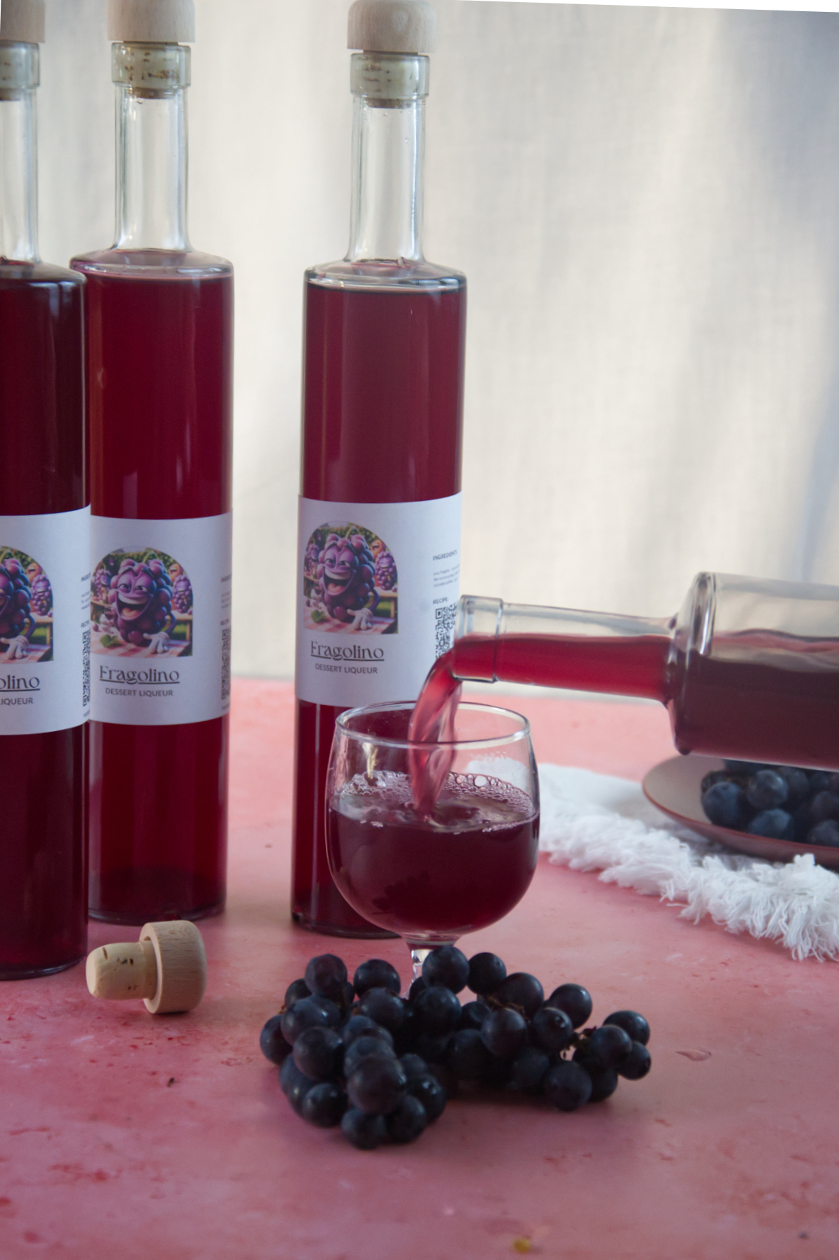 Bottiglia di liquore Fragolino rosso versata in un bicchiere con altre tre bottiglie e grappoli d'uva nera su fondo rosa materico.