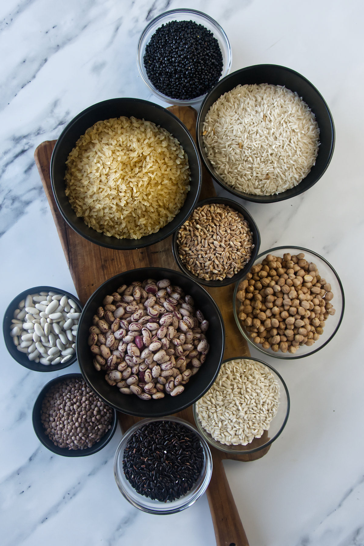 Tagliere in legno con ciotole di cereali e legumi: riso arborio, farro, ceci, lenticchie nere e fagioli borlotti.