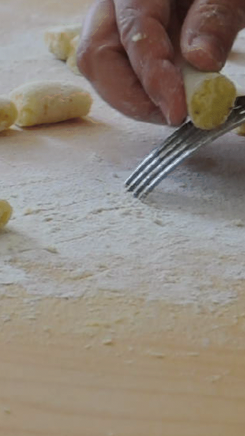 Primo piano di mani che rigano gnocchi di patate freschi sui rebbi di una forchetta su una superficie di legno infarinata.