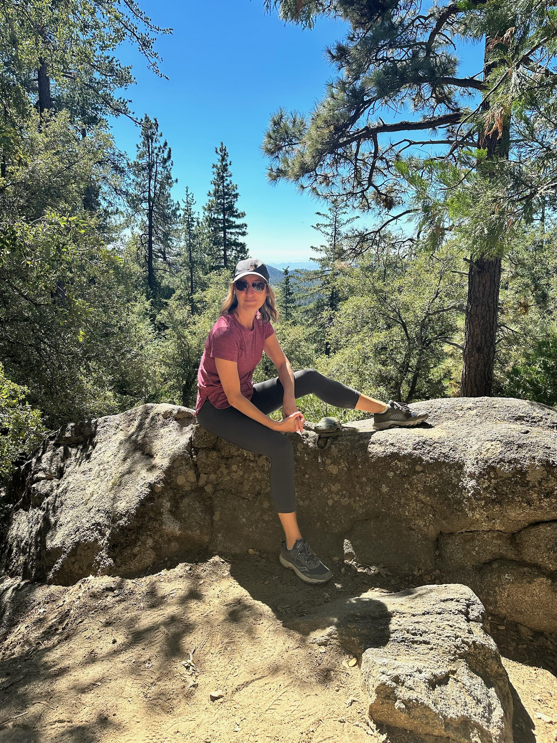 Photograph of the artist and designer in a favorite hiking trail setting, seated on a boulder with pine forest and mountains in the background. Surrounded by nature, the image portrays a source of artistic inspiration.