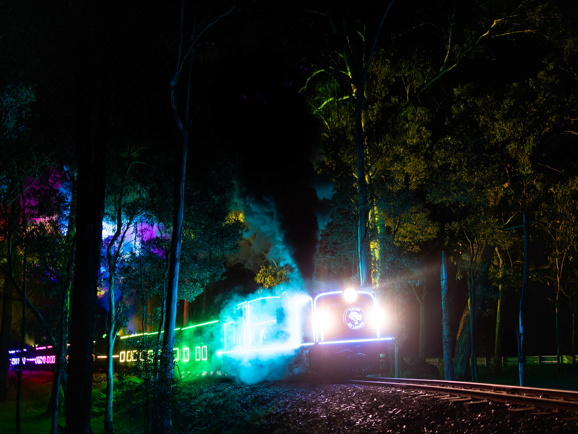 Puffing Billy's "Train of Lights" special makes a colourful exit from Fielder station as it climbs onwards to Gembrook.The Train of Lights specials, now in their second year of operation, run during the winter school holidays between Lakeside and Gembrook. The train is equipped with custom-made LED lights and projectors that cycle through a programme of colour sequences, with lights and patterns projected upwards into the forest canopy. Hauled by an ex. South African Railways garratt-type steam locomotive, the train both looks and sounds spectacular and draws crowds at every roadside location.