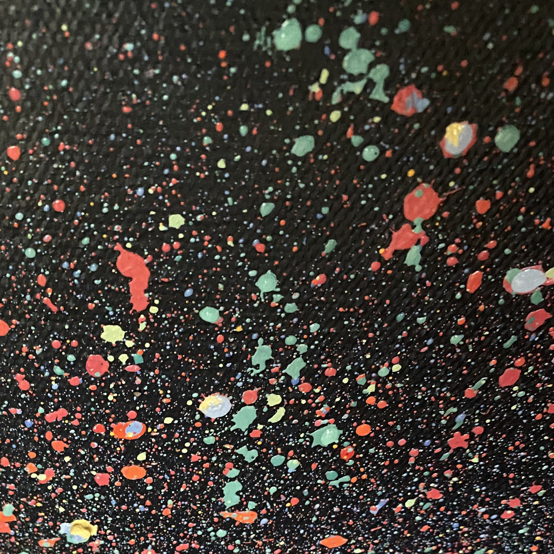 A close-up detail of a textured black canvas covered in expressive paint splatters. The composition is filled with organic, varied droplets and larger splashes of paint in bold brick red, muted sage green, and light blue. The layered speckles create a high-contrast, tactile surface with visible canvas grain, evoking a sense of chaotic cosmic dust or abstract action painting.