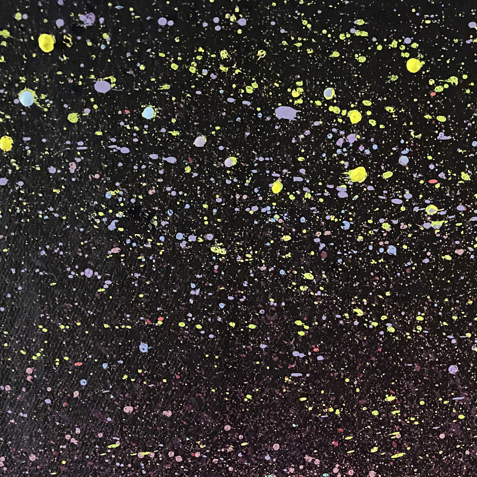 A detailed macro close-up of a painting texture showing fine splatters of paint against a dark, textured background. The splatter effect creates a dense field of tiny dots in vibrant yellow, pale violet, and soft blue, resembling a starry night sky or a distant galaxy. The lower section of the canvas transitions into subtle dark magenta and deep purple tones with finer, dust-like paint speckles.