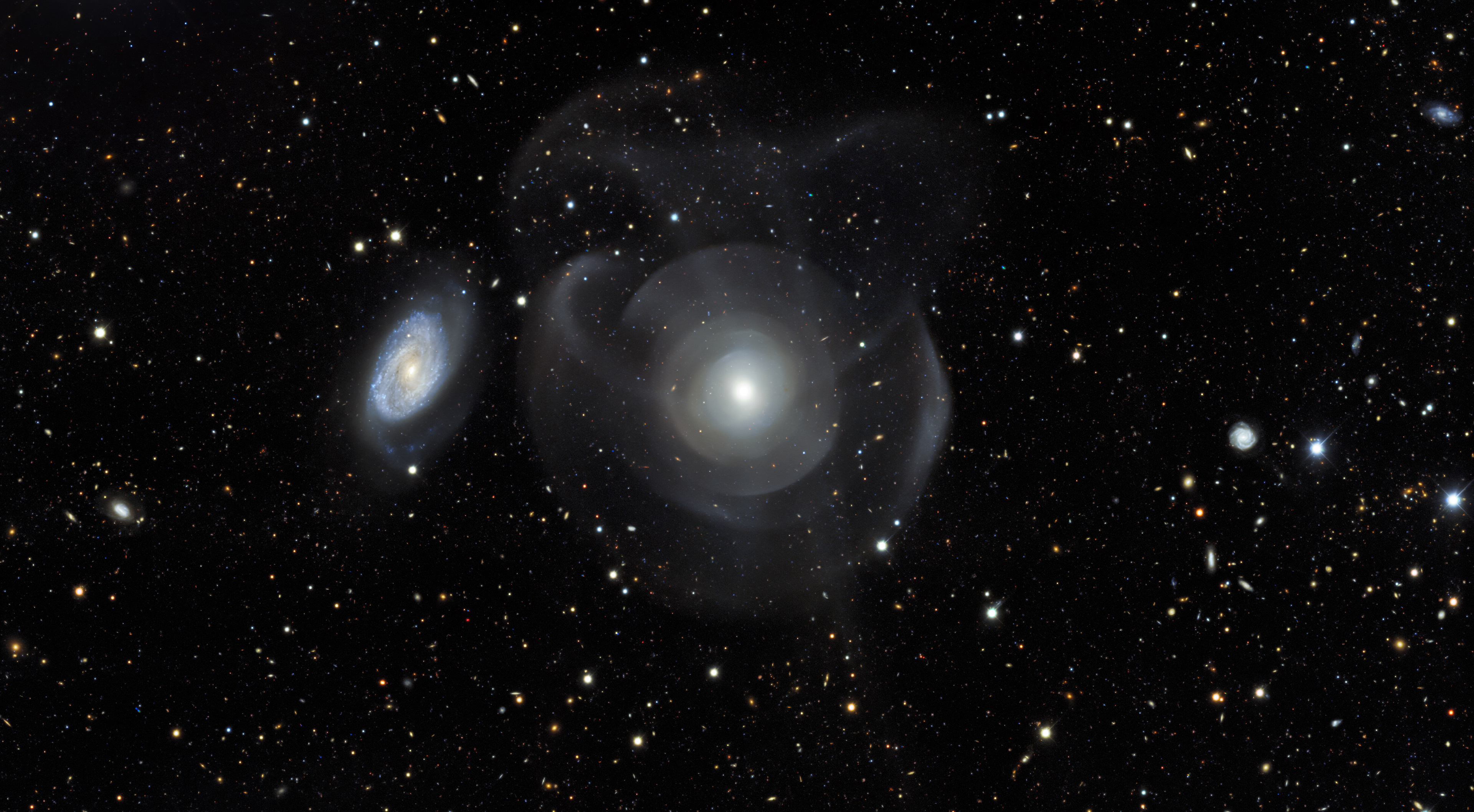 Elliptical galaxies are generally characterized by their relatively smooth appearance when compared with spiral galaxies (one of which is to the left), which have more flocculent structures interwoven with dust lanes and spiral arms. NGC 474 is at a distance of about 100 million light-years in the constellation of Pisces. This image shows unusual structures around NGC 474 characterized as tidal tails and shell-like structures made up of hundreds of millions of stars. These features are due to recent mergers (within the last billion years) or close interactions with smaller infalling dwarf galaxies.   This image is an excerpt from the Dark Energy Survey, which has released a massive, public collection of astronomical data and calibrated images from six years of work. The Dark Energy Survey is a global collaboration that includes the Department of Energy's (DOE) Fermi National Accelerator Laboratory (Fermilab), the National Center for Supercomputing Applications (NCSA), and NSF’s NOIRLab. The image was taken with the Dark Energy Camera, fabricated by DOE, on the Víctor M. Blanco 4-meter Telescope.  The quality of the survey can be appreciated by diving into the zoomable version of this wider excerpt showing a background tapestry of thousands of distant galaxies.   Credit: DES/DOE/Fermilab/NCSA & CTIO/NOIRLab/NSF/AURA Acknowledgments: Image processing: DES, Jen Miller (Gemini Observatory/NSF's NOIRLab), Travis Rector (University of Alaska Anchorage), Mahdi Zamani & Davide de Martin