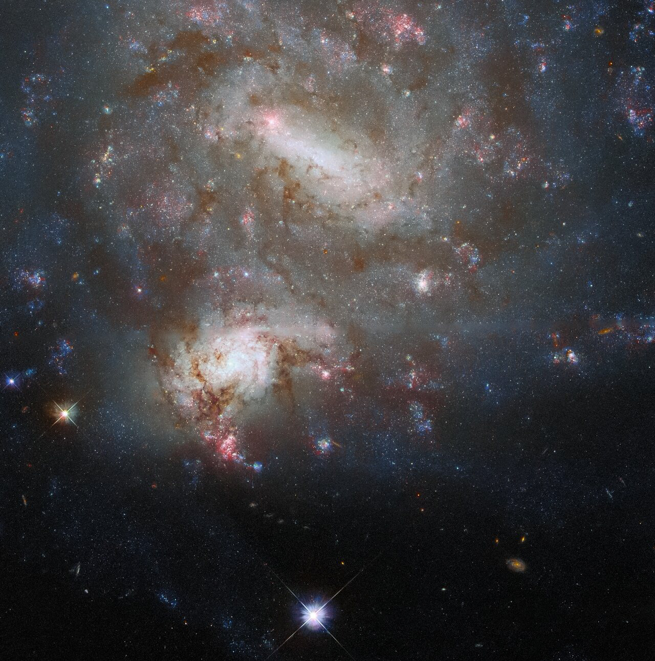 The twin galaxies NGC 4496A and NGC 4496B dominate the frame in this image from the NASA/ESA Hubble Space Telescope. Both galaxies lie in the constellation Virgo, but despite appearing side-by-side in this image they are at vastly different distances from both Earth and one another. NGC 4496A is 47 million light-years from Earth while NGC 4496B is 212 million light-years away. The enormous distances between the two galaxies mean that the two cannot interact, and they only appear to overlap owing to a chance alignment.   Chance galactic alignments such as this provide astronomers with the opportunity to delve into the distribution of dust in these galaxies. Galactic dust adds to the beauty of astronomical images — it can be seen in this image as the dark tendrils threading through both NGC 4496A and NGC 4496B — but it also complicates astronomers’ observations. Dust absorbs starlight, making stars seem dimmer and shifting their light towards longer wavelengths, a process that astronomers refer to as “reddening” (not the same thing as redshift). By carefully measuring how starlight from background galaxies is affected by dust in intervening galaxies, astronomers can map out where the dust is in the foreground galaxy’s spiral arms. The resulting “dust maps” help astronomers calibrate measurements of everything from cosmological distances to the types of stars populating galaxies.  Credit: ESA/Hubble & NASA, T. Boeker, B. Holwerda, Dark Energy Survey, DOE, FNAL/DECam, CTIO/NOIRLab/NSF/AURA, SDSS  Acknowledgement: R. Colombari