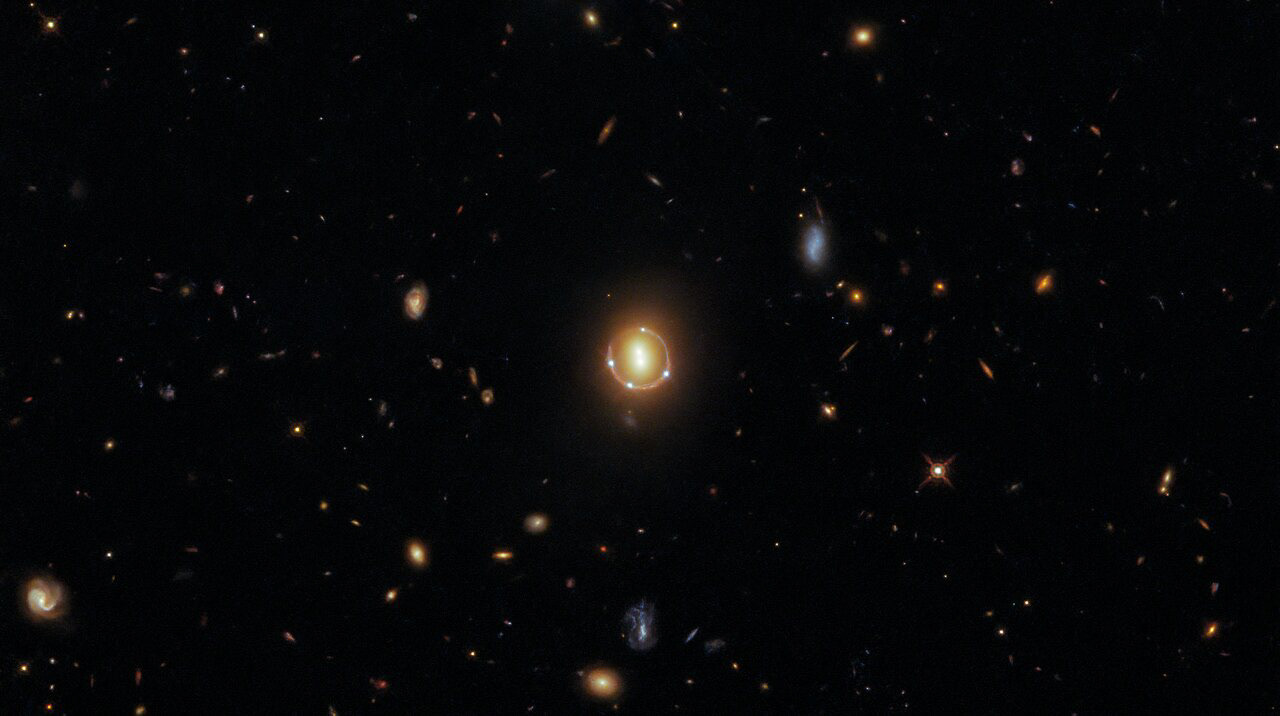 Clustered at the centre of this image are six luminous spots of light, four of them forming a circle around a central pair. Appearances can be deceiving, however, as this formation is not composed of six individual galaxies, but only three: to be precise, a pair of galaxies and one distant quasar. Hubble data also indicates that there is a seventh spot of light in the very center, which is a rare fifth image of the distant quasar. This rare phenomenon is caused by the presence of two galaxies in the foreground that act as a lens.  These galaxies were imaged in spectacular detail by Hubble’s Wide Field Camera 3 (WFC3), which was installed on Hubble in 2009 during Hubble Servicing Mission 4, Hubble’s final servicing mission. The WFC3 was intended to operate until 2014, but 12 years after it was installed it continues to provide both top-quality data and fantastic images, such as this one.   The central pair of galaxies in this image are genuinely two separate galaxies. The four bright points circling them, and the fainter one in the very center, are actually five separate images of a single quasar (known as 2M1310-1714), an extremely luminous but distant object. The reason behind this “seeing quintuple” effect is a phenomenon known as gravitational lensing. Gravitational lensing occurs when a celestial object with an enormous amount of mass — such as a pair of galaxies — causes the fabric of space to warp such that the light travelling through that space from a distant object is bent and magnified sufficiently that humans here on Earth can observe multiple magnified images of the far-away source. The quasar in this image actually lies further away from Earth than the pair of galaxies. The light from the quasar has been bent around the galaxy pair because of their enormous mass, giving the incredible appearance that the galaxy pair are surrounded by four quasars  — whereas in reality, a single quasar lies far beyond them!  Links Video of Seeing Quintuple Credit: ESA/Hubble & NASA, T. Treu  Acknowledgment: J. Schmidt