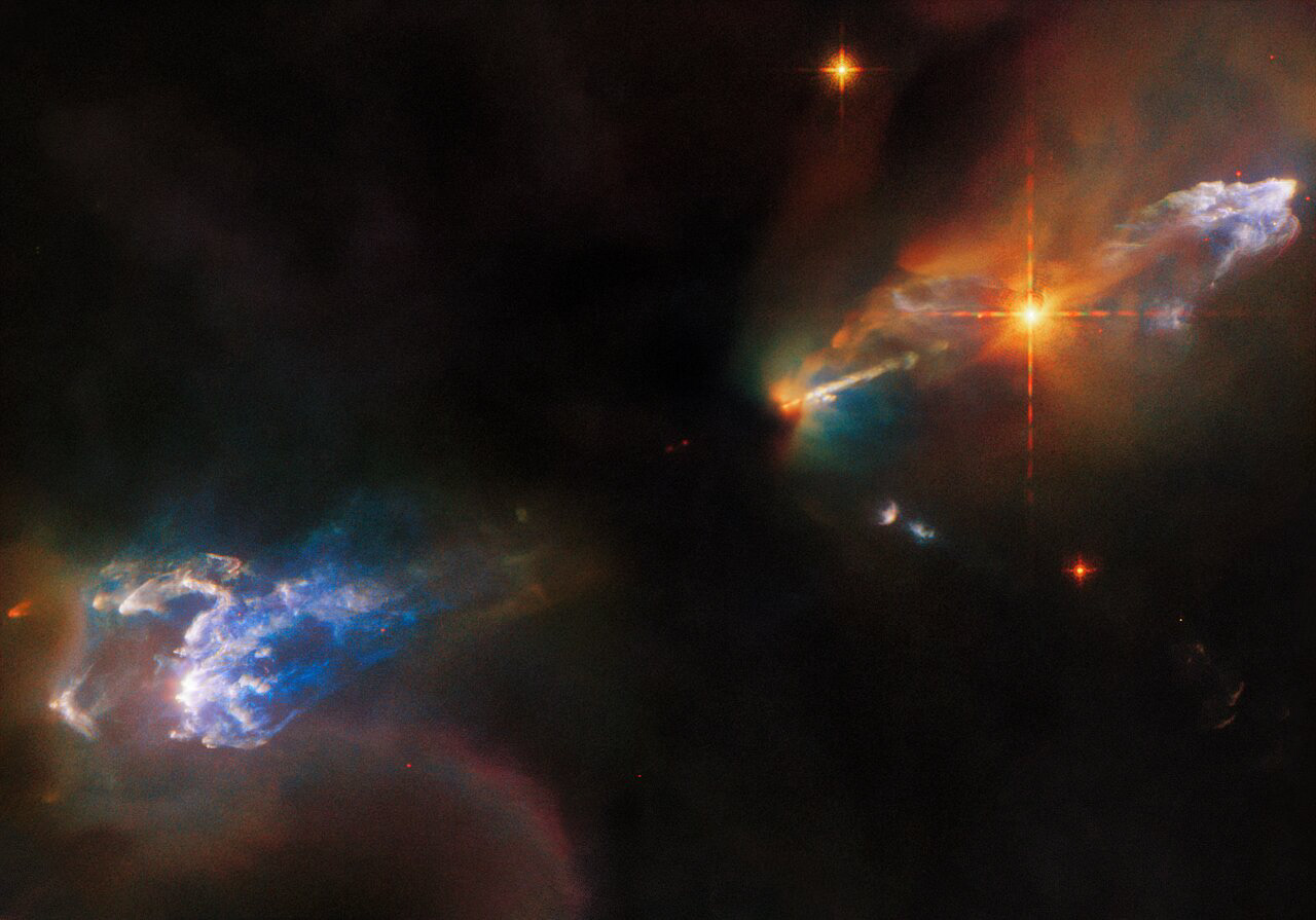 The lives of newborn stars are tempestuous, as this image of the Herbig–Haro objects HH 1 and HH 2 from the NASA/ESA Hubble Space Telescope depicts. Both objects are in the constellation Orion and lie around 1250 light-years from Earth. HH 1 is the luminous cloud above the bright star in the upper right of this image, and HH 2 is the cloud in the bottom left. While both Herbig–Haro objects are visible, the young star system responsible for their creation is lurking out of sight, swaddled in the thick clouds of dust at the centre of this image. However, an outflow of gas from one of these stars can be seen streaming out from the central dark cloud as a bright jet. Meanwhile, the bright star between that jet and the HH 1 cloud was once thought to be the source of these jets, but it is now known to be an unrelated double star that formed nearby.  Herbig–Haro objects are glowing clumps found around some newborn stars, and are created when jets of gas thrown outwards from these young stars collide with surrounding gas and dust at incredibly high speeds. In 2002 Hubble observations revealed that parts of HH 1 are moving at more than 400 kilometres per second!  This scene from a turbulent stellar nursery was captured with Hubble’s Wide Field Camera 3 using 11 different filters at infrared, visible, and ultraviolet wavelengths. Each of these filters is sensitive to just a small slice of the electromagnetic spectrum, and they allow astronomers to pinpoint interesting processes that emit light at specific wavelengths.  In the case of HH 1/2, two groups of astronomers requested Hubble observations for two different studies. The first delved into the structure and motion of the Herbig–Haro objects visible in this image, giving astronomers a better understanding of the physical processes occurring when outflows from young stars collide with surrounding gas and dust. The second study instead investigated the outflows themselves to lay the groundwork for future observations with the NASA/ESA/CSA James Webb Space Telescope. Webb, with its ability to peer past the clouds of dust enveloping young stars, will revolutionise the study of outflows from young stars.  [Image description: Two wispy, gaseous clouds occupy the corners of this image, HH 1 in the upper right, and HH 2 in the lower left. Both are light blue and surrounded by dimmer multi-coloured clouds, while the background is dark black due to dense gas. A very bright orange star lies just to the lower left of HH 1, and beyond that star is a narrow jet, emerging from the dark centre of the field.]  Links Video of Multiwavelength View of a Turbulent Stellar Nursery Credit: ESA/Hubble & NASA, B. Reipurth, B. Nisini