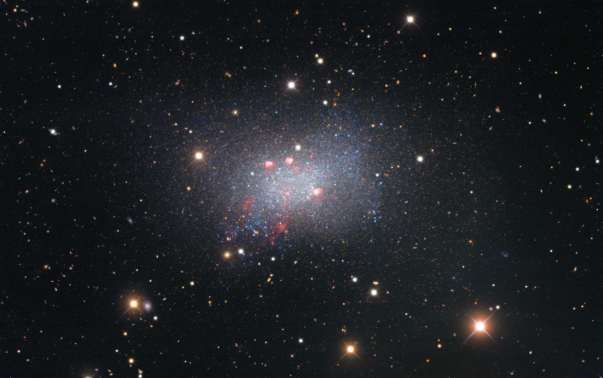 Sextans B is an irregular dwarf galaxy, meaning that it is irregularly shaped and smaller than our galaxy, the Milky Way. It lies around 4.5 million light-years from Earth and is located in the constellation Sextans in the southern sky. Captured with the Nicholas U. Mayall 4-meter Telescope, this image of Sextans B features red-colored star-forming regions near the galaxy’s center. Surrounding the galaxy are several bright stars that are located much closer to us in our galaxy, identified by the crisscross patterns created by light interacting with the structure of the telescope, as well as numerous fuzzy-looking background galaxies that appear small because they are much farther away than Sextans B.  Credit: KPNO/NOIRLab/NSF/AURA Data obtained and processed by: P. Massey (Lowell Obs.), G. Jacoby, K. Olsen, & C. Smith (AURA/NSF) Image processing: T.A. Rector (University of Alaska Anchorage/NSF’s NOIRLab), M. Zamani (NSF’s NOIRLab) & D. de Martin (NSF’s NOIRLab)