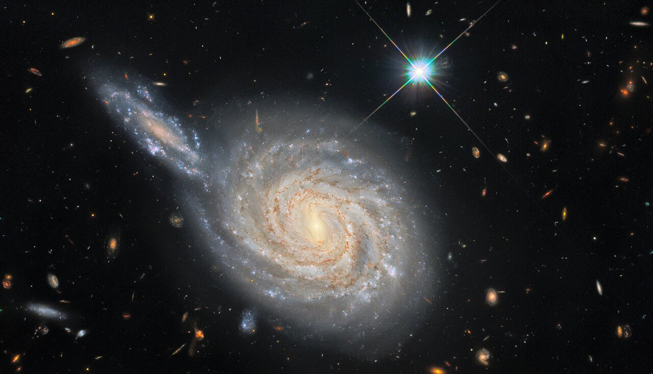 This image from the NASA/ESA Hubble Space Telescope captures the spiral galaxy NGC 105, which lies roughly 215 million light-years away in the constellation Pisces. While it looks like NGC 105 is plunging edge-on into a collision with a neighbouring galaxy, this is just the result of the chance alignment of the two objects in the night sky. NGC 105’s elongated neighbour is actually far more distant and remains relatively unknown to astronomers. These misleading conjunctions occur frequently in astronomy — for example, the stars in constellations are at vastly different distances from Earth, and only appear to form patterns thanks to the chance alignment of their component stars.  The Wide Field Camera 3 observations in this image are from a vast collection of Hubble measurements examining nearby galaxies which contain two fascinating astronomical phenomena — Cepheid variables and cataclysmic supernova explosions. Whilst these two phenomena may appear to be unrelated — one is a peculiar class of pulsating stars and the other is the explosion caused by the catastrophic final throes of a massive star’s life — they are both used by astronomers for a very particular purpose: measuring the vast distances to astronomical objects. Both Cepheids and supernovae have very predictable luminosities, meaning that astronomers can tell precisely how bright they are. By measuring how bright they appear when observed from Earth, these “standard candles” can provide reliable distance measurements. NGC 105 contains both supernovae and Cepheid variables, giving astronomers a valuable opportunity to calibrate the two distance measurement techniques against one another.  Astronomers recently carefully analysed the distances to a sample of galaxies including NGC 105 to measure how fast the Universe is expanding — a value known as the Hubble constant. Their results don’t agree with the predictions of the most widely-accepted cosmological model, and their analysis shows that there is only a 1-in-a-million chance that this discrepancy was caused by measurement errors. This discrepancy between galaxy measurements and cosmological predictions has been a long-standing source of consternation for astronomers, and these recent findings provide persuasive new evidence that something is either wrong or lacking in our standard model of cosmology.  Credit: ESA/Hubble & NASA, D. Jones, A. Riess et al. Acknowledgement: R. Colombari