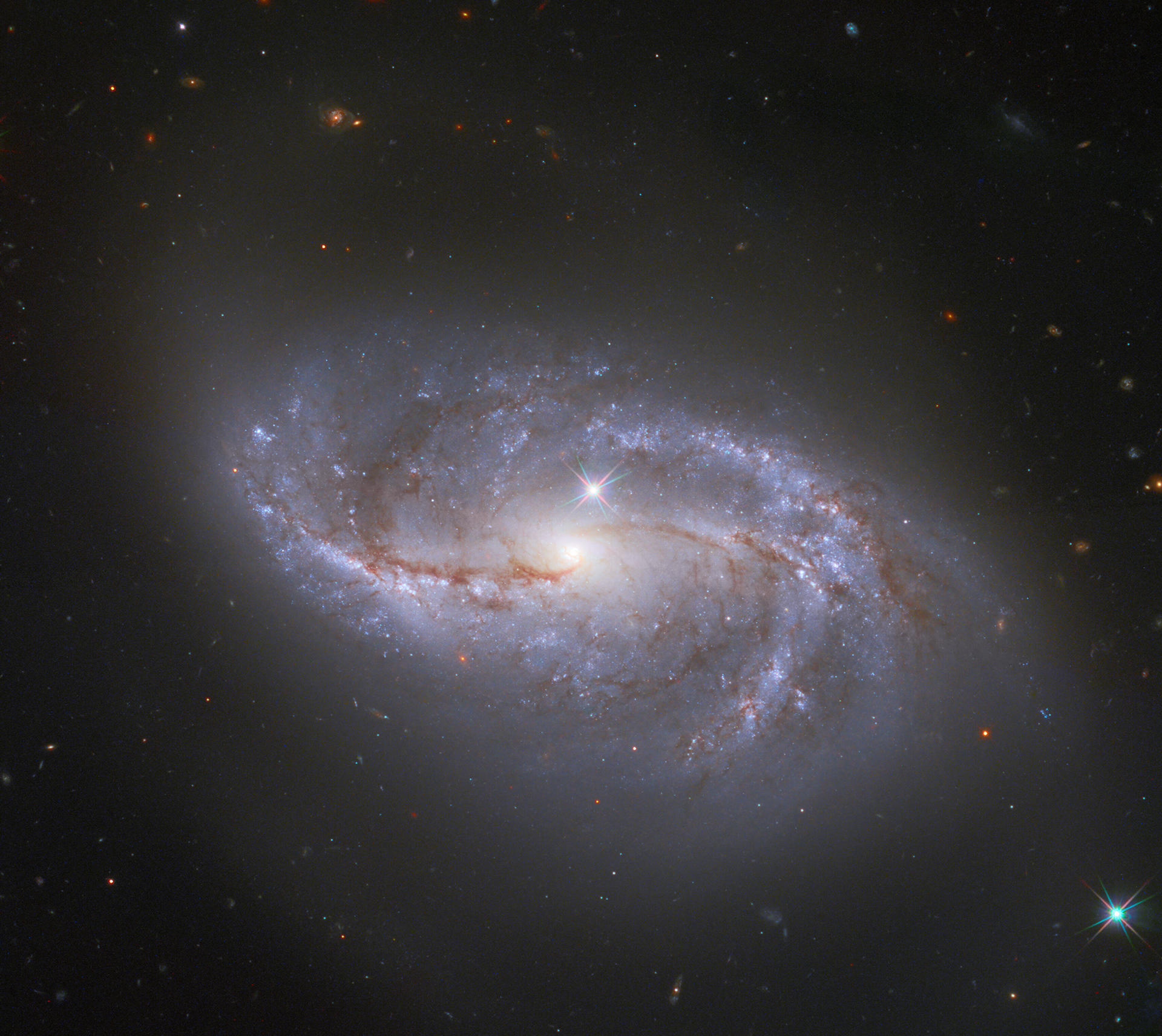 One Amongst Millions / Looking deep into the Universe, the NASA/ESA Hubble Space Telescope catches a passing glimpse of the numerous arm-like structures that sweep around this barred spiral galaxy, known as NGC 2608. Appearing as a slightly stretched, smaller version of our Milky Way, the peppered blue and red spiral arms are anchored together by the prominent horizontal central bar of the galaxy.  In Hubble photos, bright Milky Way stars will sometimes appear as pinpoints of light with prominent lens flares. A star with these features is seen in the lower right corner of the image, and another can be spotted just above the pale centre of the galaxy. The majority of the fainter points around NGC 2608, however, lack these features, and upon closer inspection they are revealed to be thousands of distant galaxies. NGC 2608 is just one among an uncountable number of kindred structures.  Similar expanses of galaxies can be observed in other Hubble images such as the Hubble Deep Field which recorded over 3000 galaxies in one field of view.  Credit: ESA/Hubble & NASA, A. Riess et al.