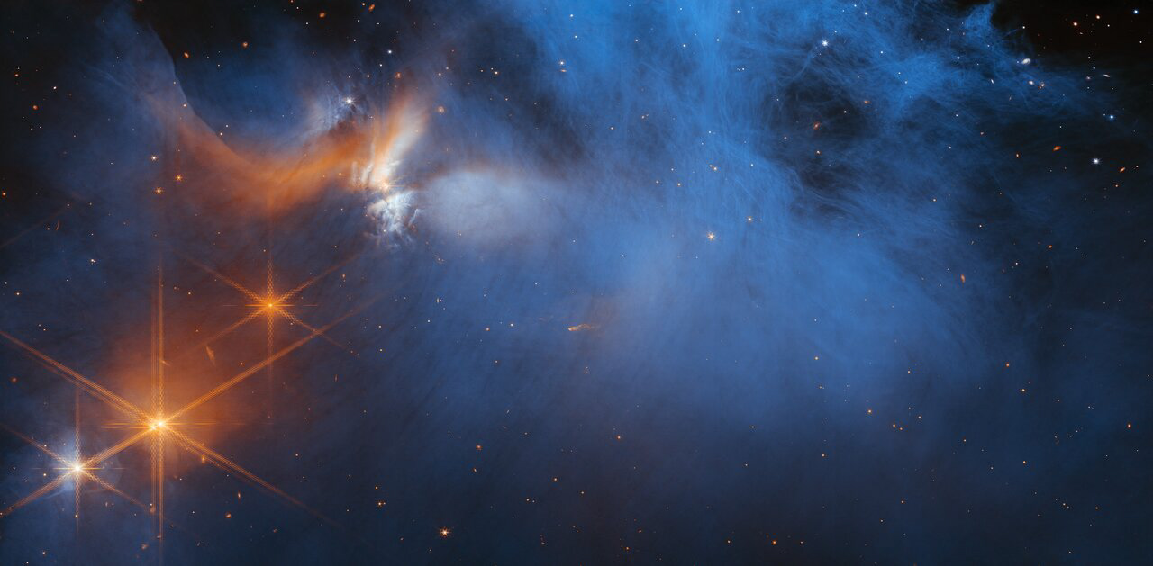 This image by the NASA/ESA/CSA James Webb Space Telescope’s Near-InfraRed Camera (NIRCam) features the central region of the Chameleon I dark molecular cloud, which resides 630 light years away. The cold, wispy cloud material (blue, centre) is illuminated in the infrared by the glow of the young, outflowing protostar Ced 110 IRS 4 (orange, upper left). The light from numerous background stars, seen as orange dots behind the cloud, can be used to detect ices in the cloud, which absorb the starlight passing through them.   An international team of astronomers has reported the discovery of diverse ices in the darkest, coldest regions of a molecular cloud measured to date by studying this region. This result allows astronomers to examine the simple icy molecules that will be incorporated into future exoplanets, while opening a new window on the origin of more complex molecules that are the first step in the creation of the building blocks of life.  [Image Description: A large, dark cloud is contained within the frame. In its top half it is textured like smoke and has wispy gaps, while at the bottom and at the sides it fades gradually out of view. On the left are several orange stars: three each with six large spikes, and one behind the cloud which colours it pale blue and orange. Many tiny stars are visible, and the background is black.]  Credit: NASA, ESA, CSA, and M. Zamani (ESA/Webb); Science: F. Sun (Steward Observatory), Z. Smith (Open University), and the Ice Age ERS Team.