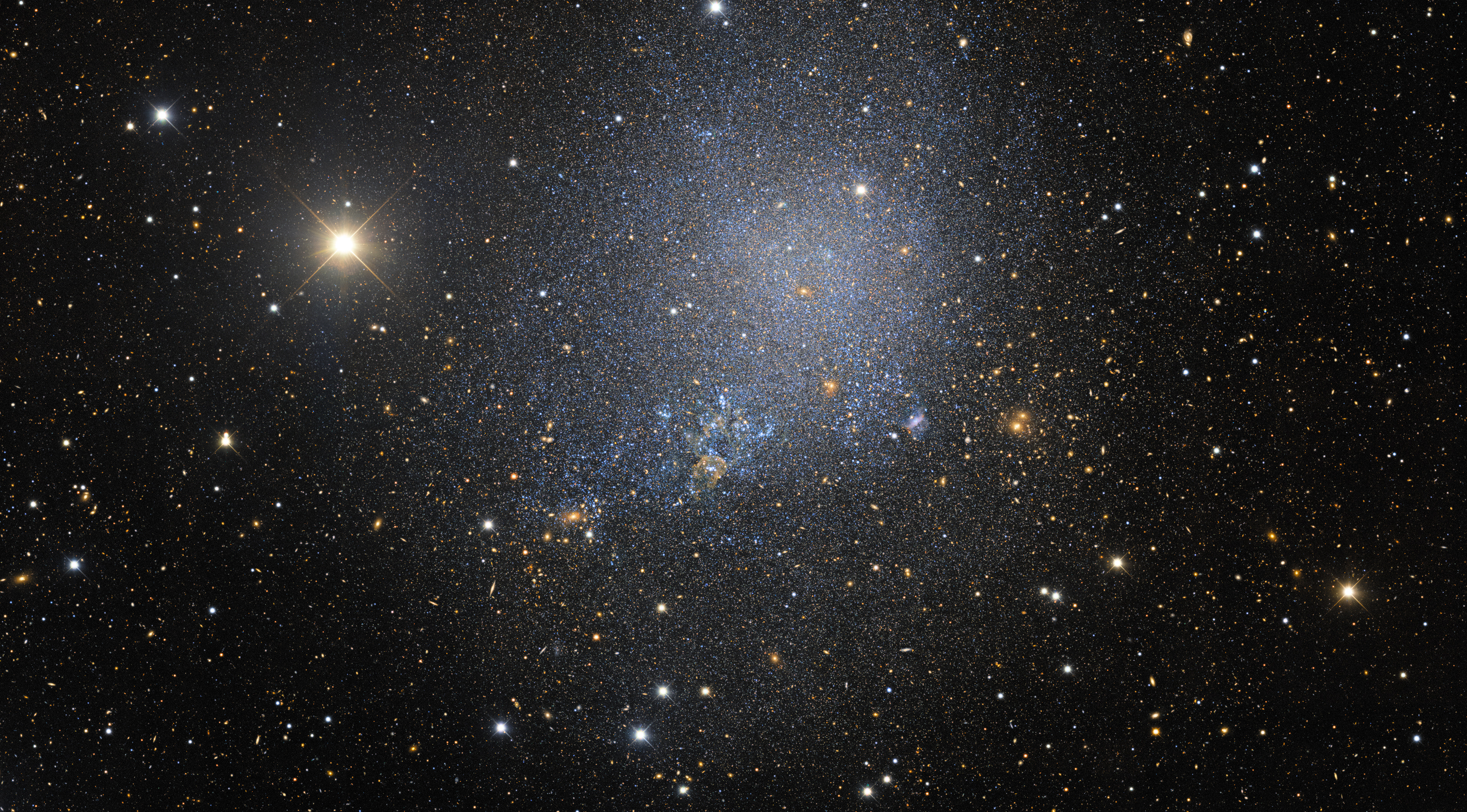 The irregular dwarf galaxy IC 1613 contains some 100 million stars and is a member of our Local Group of galaxies, which also includes our Milky Way, the Andromeda spiral galaxy, and the Magellanic Clouds. It is at a distance of 2.4 million light-years and contains several examples of Cepheid variable stars — key calibrators of the cosmic distance ladder. The bulk of its stars were formed about 7 billion years ago and it does not appear to be undergoing star formation at the present day, unlike other very active dwarf irregulars such as the Large and Small Magellanic clouds.  To the lower right of IC 1613, one may view a background galaxy cluster (several hundred times more distant than IC 1613) consisting of dozens of orange-yellow blobs, centered on a pair of giant cluster elliptical galaxies. To the left of the irregular galaxy is a bright, sixth magnitude, foreground, Milky Way star in the constellation of Cetus the Whale, identified here as a star by its sharp diffraction spikes radiating at 45-degree angles.   This image is an excerpt from the Dark Energy Survey, which has released a massive, public collection of astronomical data and calibrated images from six years of work. The Dark Energy Survey is a global collaboration that includes the Department of Energy's (DOE) Fermi National Accelerator Laboratory (Fermilab), the National Center for Supercomputing Applications (NCSA), and NSF’s NOIRLab. The image was taken with the Dark Energy Camera, fabricated by DOE, on the Víctor M. Blanco 4-meter Telescope.  The immensity and depth of the survey can be appreciated by diving into the zoomable version of this wider excerpt.   Credit: DES/DOE/Fermilab/NCSA & CTIO/NOIRLab/NSF/AURA Acknowledgments: Image processing: DES, Jen Miller (Gemini Observatory/NSF's NOIRLab), Travis Rector (University of Alaska Anchorage), Mahdi Zamani & Davide de Martin